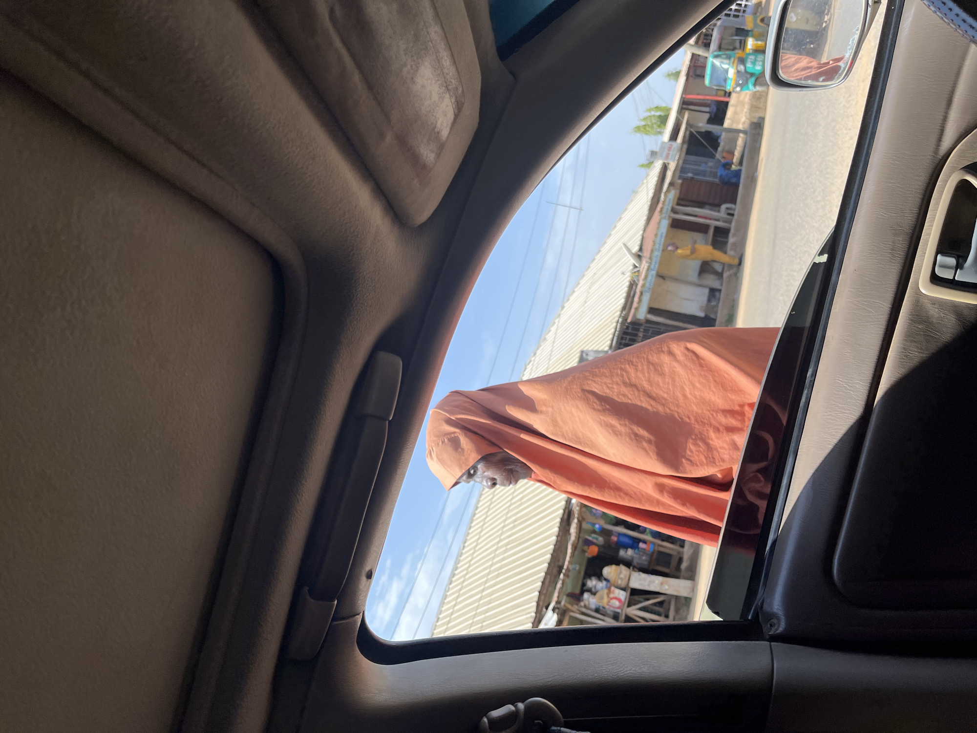 The image, taken from inside a car through a partially opened window, shows a woman in a light-orange, full-body covering standing outside. The location appears to be a street in Barakin Akawo, Nigeria, based on the urban setting and nearby architecture. There are structures with corrugated metal roofs and various merchandise displayed outside, typical of commercial roadside establishments. Other people and vehicles (possibly auto rickshaws) are visible further down the street. The focus is on the woman at the car window.