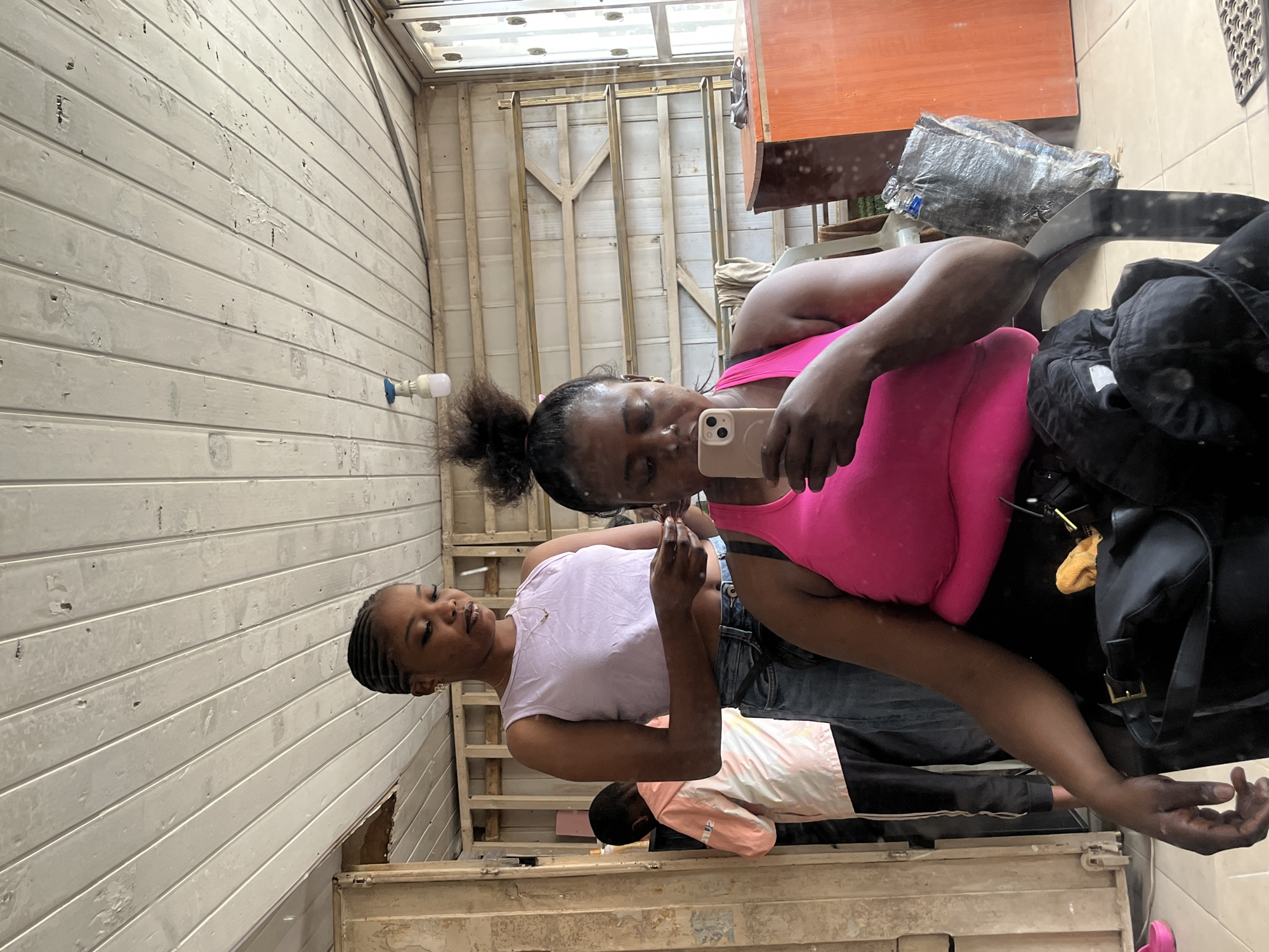 The image depicts an indoor scene with a woman in the foreground taking a selfie with her phone. She is wearing a bright pink tank top and black bottoms. To her left, another woman in a light purple crop top and jeans is looking down, possibly at her hands. Further to the left, a child in a light pink hooded jacket and dark pants is partially visible. The environment appears to be an unfinished or industrial space, with exposed wooden beams on the ceiling and walls. A desk with an orange surface is visible on the right side of the image. The overall lighting is natural, coming from the right side of the frame. The scene appears to be located in Abuja, Nigeria.