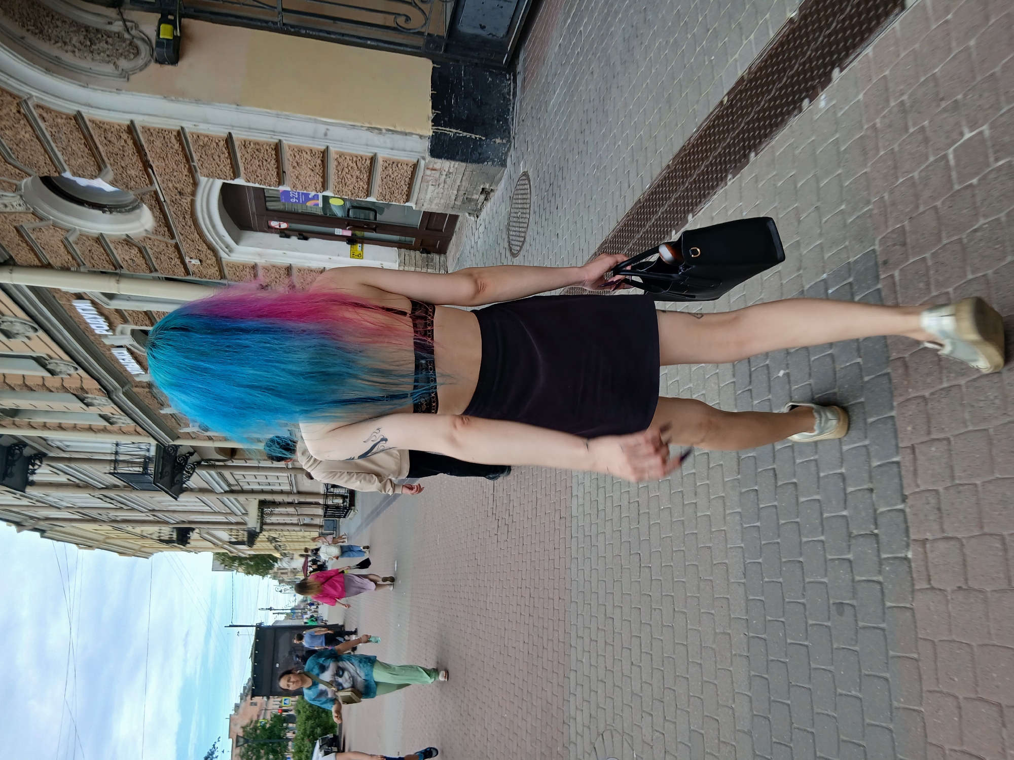 A young woman with vibrant blue and pink hair walks away from the camera on a cobblestone sidewalk in front of a building. She is wearing a black skirt and a bra. She carries a black handbag in her right hand. Other pedestrians are visible in the background on the sidewalk, and the building facade features arched windows and ornate details. The sky is overcast.