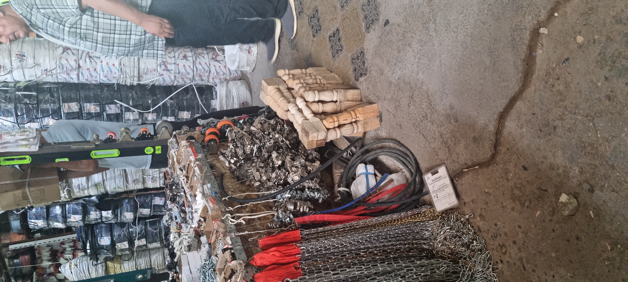The image depicts an indoor market stall, likely in Bokhtar, Tajikistan. A man wearing a plaid shirt and dark pants stands to the right, facing away from the camera. The stall is filled with various hardware and supplies. In the foreground, to the left, are several chains and red straps. To the right of the chains is a pile of metal hinges, and further to the right are stacks of wooden balusters. In the background, numerous rolls of wire and cable are stacked high. The floor is concrete with a patterned design. The overall scene suggests a retail environment for building materials or hardware.