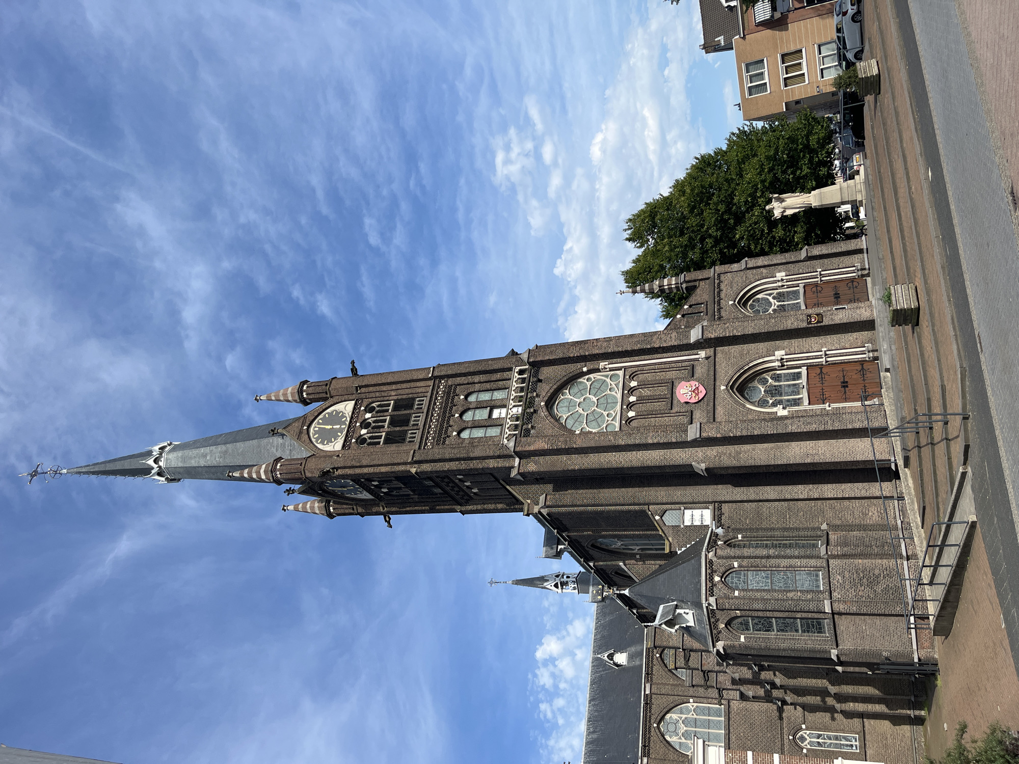 A brick church with a tall, dark spire dominates the frame under a bright blue sky with scattered white clouds. The church has multiple arched windows, some with stained glass, and a prominent clock face on its tower. The entrance to the church is up a set of stairs, with a ramp alongside. To the right of the church, a statue of a robed figure stands in front of large green trees. Beyond the trees, modern buildings, one with a light brown facade, are visible. Several cars are parked on the street in front of the buildings. The ground in the foreground is paved with brick.