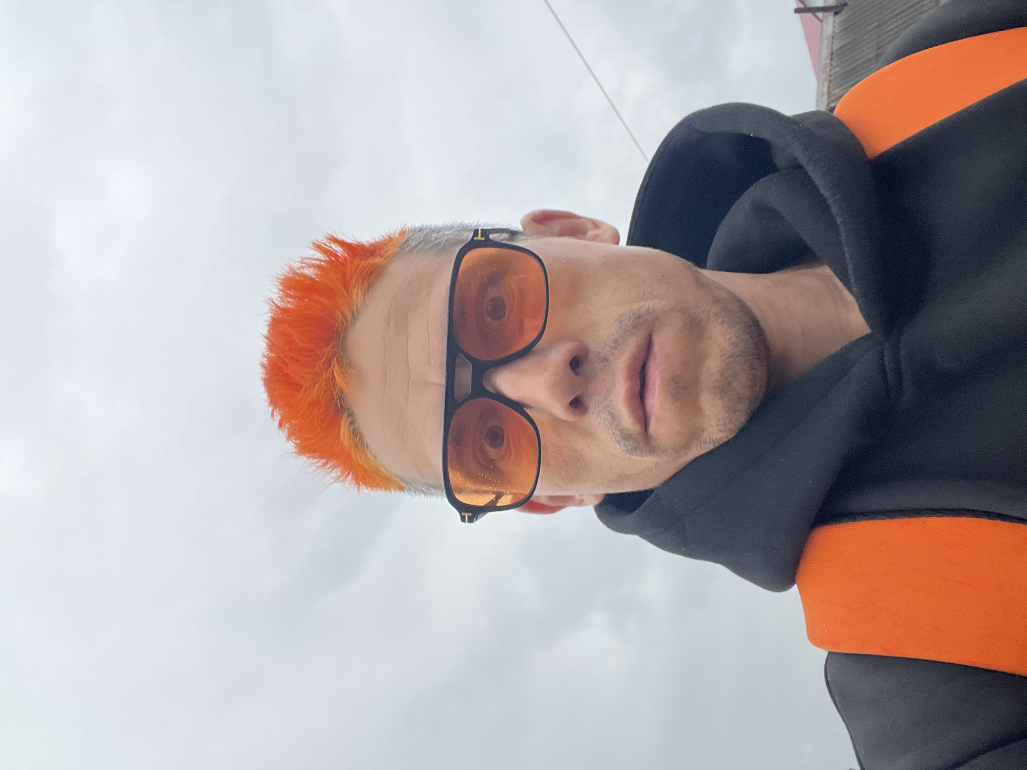 A low-angle outdoor selfie shows a person with brightly orange hair, black-framed sunglasses with orange lenses, and a black hoodie with orange trim. The sky is overcast. The person is possibly in Chaikovsky, Russia, but there is no specific detail in the image to confirm that location.