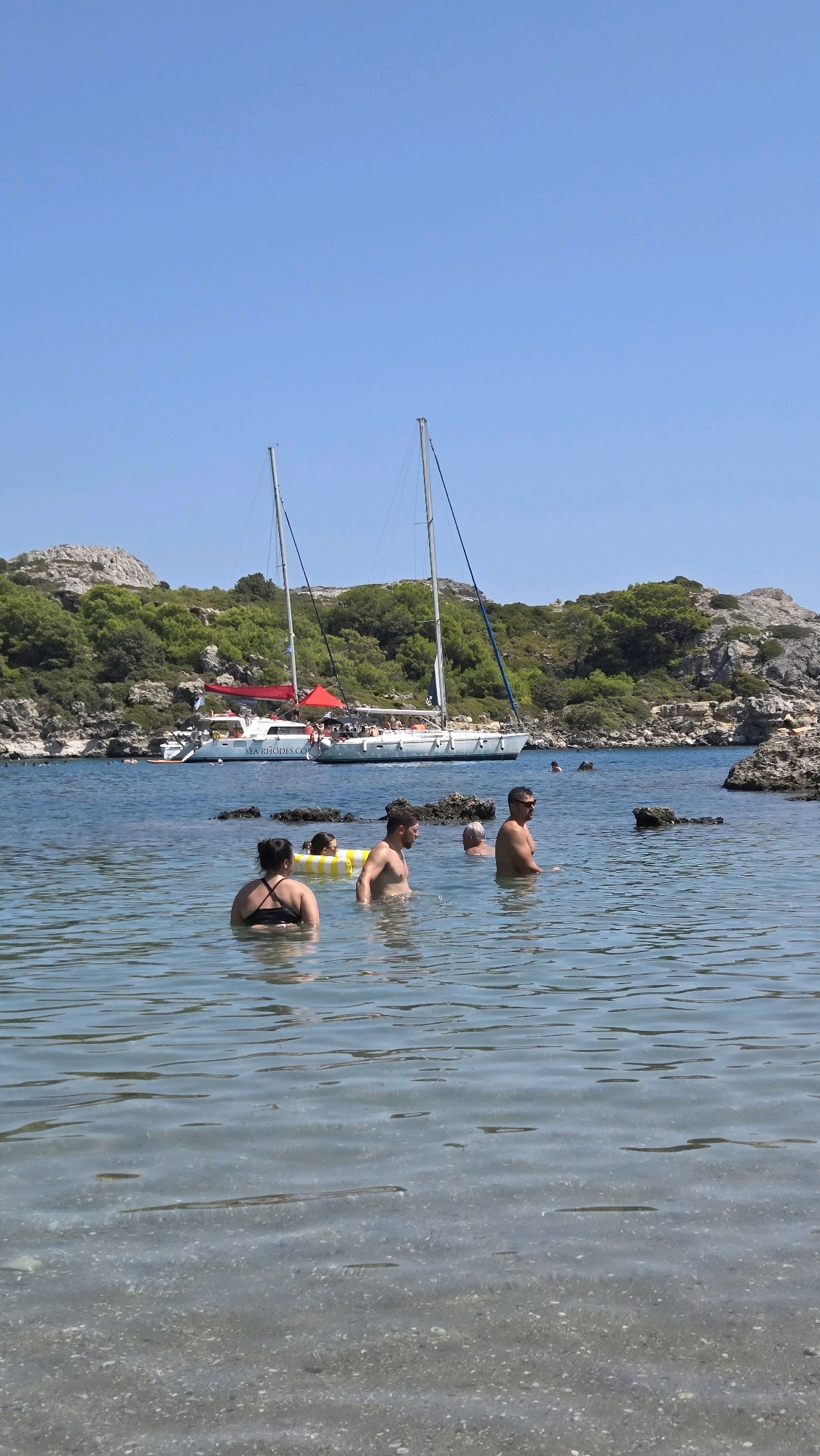 The image depicts a sunny day at a coastal location in Rhodes, Greece, likely a bay or cove. In the foreground, several individuals are wading in clear, shallow water. To the left, a woman wearing a black bikini is partially submerged. Next to her, a child wearing a yellow and white striped inflatable ring is visible. A man with a bare chest is in the center, looking to his right. Another man, also bare-chested, is further to the right, wearing sunglasses. In the middle ground, a white catamaran with red accents and the text "SEA RHODES.COM" on its side is anchored. The catamaran has multiple passengers on deck and two tall masts. Behind the catamaran, a rocky, tree-covered hillside forms the backdrop. The water is calm with visible ripples, and the seabed is rocky and pebbly. The sky is clear blue.