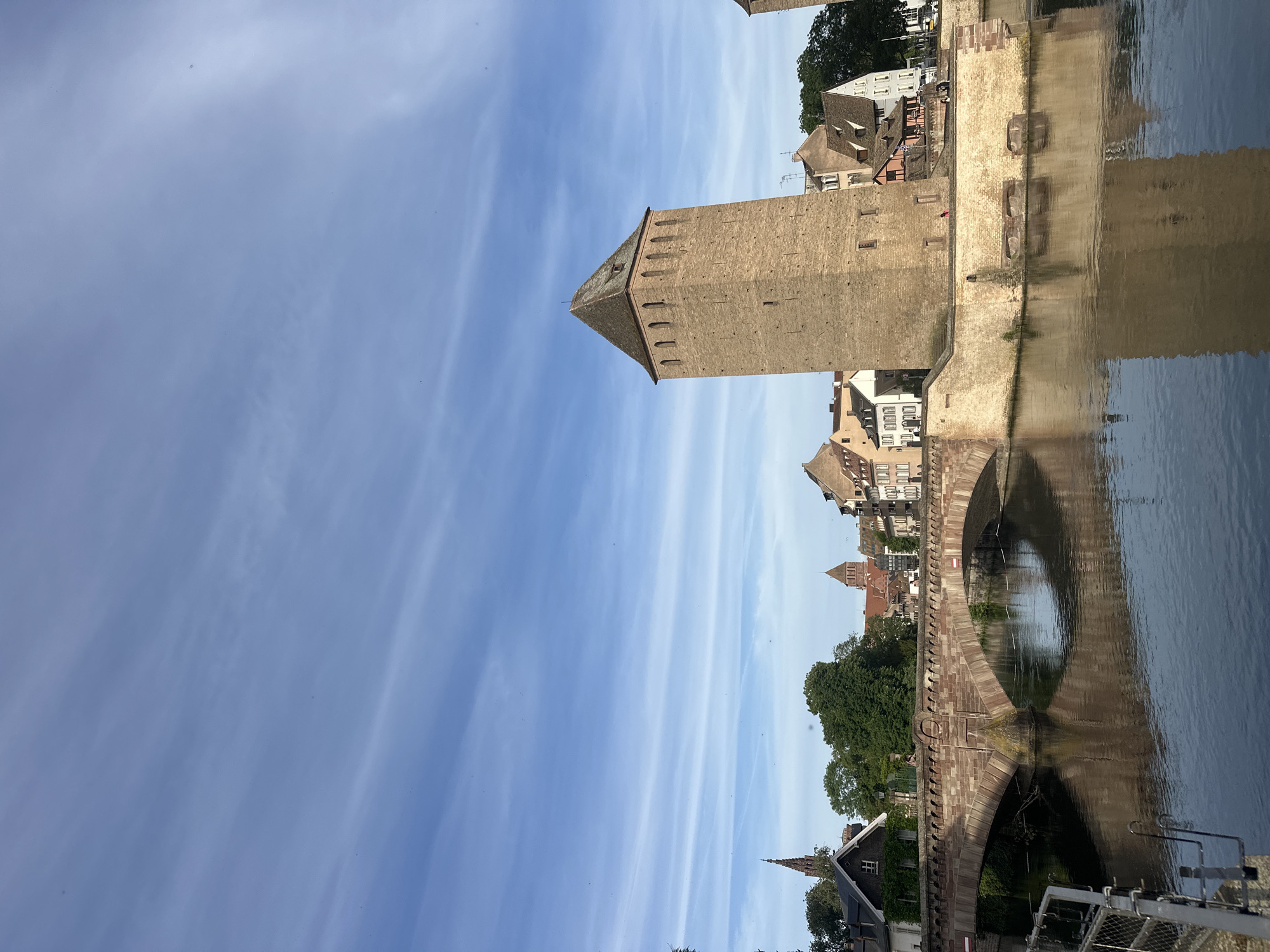 The image captures the Ponts Couverts in Strasbourg, France. The Ponts Couverts are a set of three bridges and four towers that were part of the city's defensive fortifications. The towers are built of brick and have steep, pyramid-shaped roofs. The bridges span the Ill River, and their arches are reflected in the water. Buildings line the riverbanks. The sky is blue with cirrus clouds.