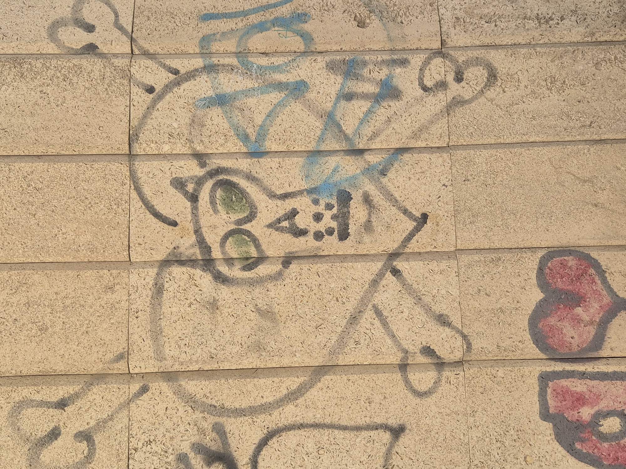The image displays graffiti on a wall composed of beige, rectangular textured panels. The graffiti includes a stylized skull with green eyes, a nose, and dots for teeth, framed by a heart shape and crossbones. There are also partial shapes of hearts with red infill at the bottom of the image, as well as blue spray paint markings that appear to be letters or abstract shapes overlaying the central skull design. The scene is lit by natural sunlight, casting shadows that highlight the textured surface of the wall. The graffiti is located on an exterior wall in Kokshetau, Kazakhstan.