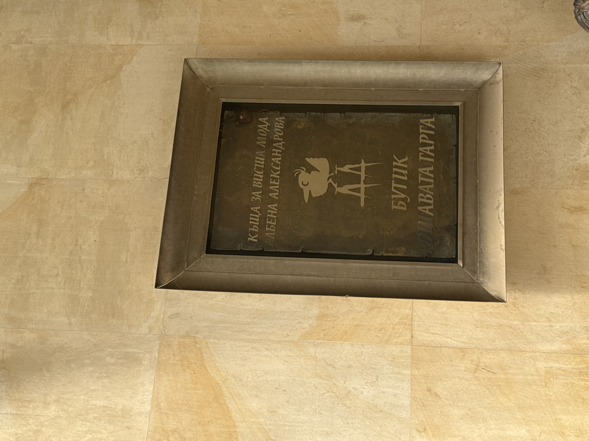 The image displays a rectangular metal plaque, possibly a floor sign, embedded within light-colored, sandy-toned floor tiles. The plaque is framed by a slightly raised, metallic border. The overall setting appears to be an indoor space, likely a commercial establishment, given the nature of the text on the plaque.
The plaque itself is made of a dark, aged metal with lighter, embossed or engraved text and a central emblem. The text is in Cyrillic, oriented vertically along the longer sides of the rectangle, and reads from top to bottom.
On the left side, the text says:
"КЪЩА ЗА ВИСША МОДА" (KUSHTA ZA VISHA MODA), which translates to "House for High Fashion".
Below that is "АЛБЕНА АЛЕКСАНДРОВА" (ALBENA ALEKSANDROVA), identifying a person, likely the designer or owner.
In the center of the plaque is a stylized emblem featuring what appears to be a bird, possibly a phoenix or a rooster, above two interlocking or stylized 'A' letters.
On the right side, the text reads:
"БУТИК" (BUTIK), meaning "Boutique".
Below that is "ПЛАВАТА ГАРТА" (PLAVATA GARTA), which is likely the name of the boutique or brand.
The floor tiles are large and rectangular, showing natural variations in color and texture, suggesting a stone-like material. No people or activities are visible, and the time of day is not ascertainable from the indoor lighting. The plaque's content directly indicates a high-fashion boutique run by Albena Alexandrova in Sofia, Bulgaria.