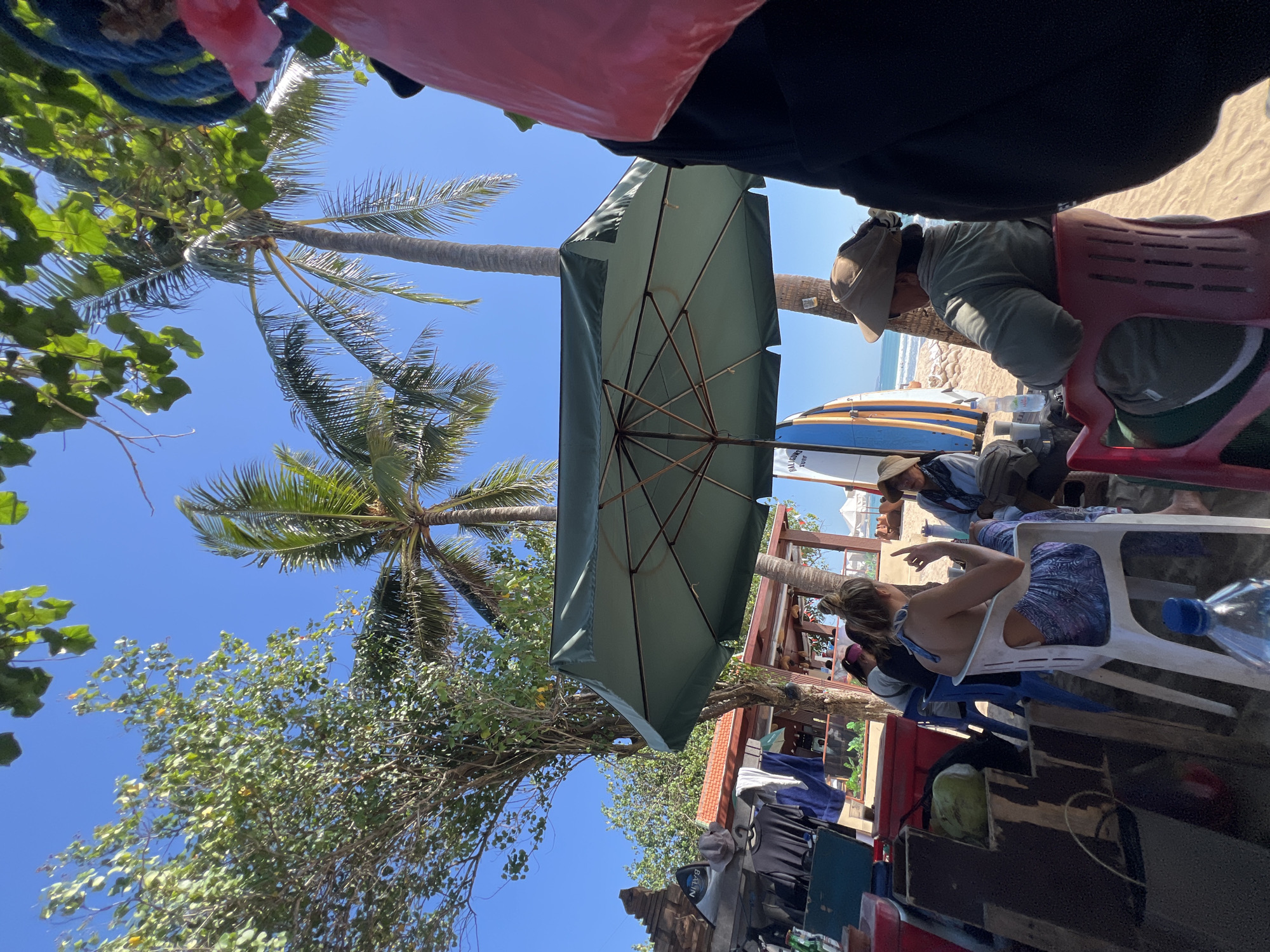 In Kuta, Indonesia, under the shade of a large green umbrella, three people are seated on chairs on a sandy beach. A woman with blonde hair pulled into a bun is wearing a blue and white patterned skirt and a bikini top, facing left and gesturing with her hand. To her right, a person wearing a hat and a green jacket sits facing away from the camera. Another individual, partially visible, is seated to the left of the woman with blonde hair. In the background, palm trees, surfboards, and beach umbrellas are visible. Various items, including clothing and a green cooler, are arranged on a wooden structure to the left of the frame. The scene appears to be a casual gathering at a beachside establishment.
