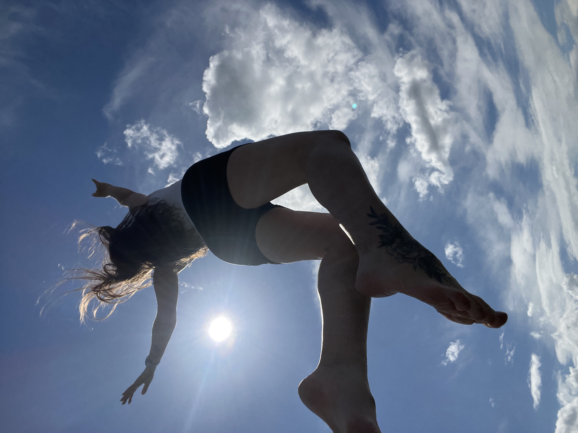 A low-angle shot captures a person in mid-air, silhouetted against a bright sun and a blue sky dotted with white clouds. The individual is wearing dark shorts and a light-colored top, with their arms outstretched and legs extended. Their hair is long and appears to be flowing in the wind. Visible tattoos can be seen on one of the person's feet. The sun is positioned in the upper left quadrant of the frame, creating a lens flare. The overall impression is one of freedom and motion. The location is given as Borychivka, Ukraine.