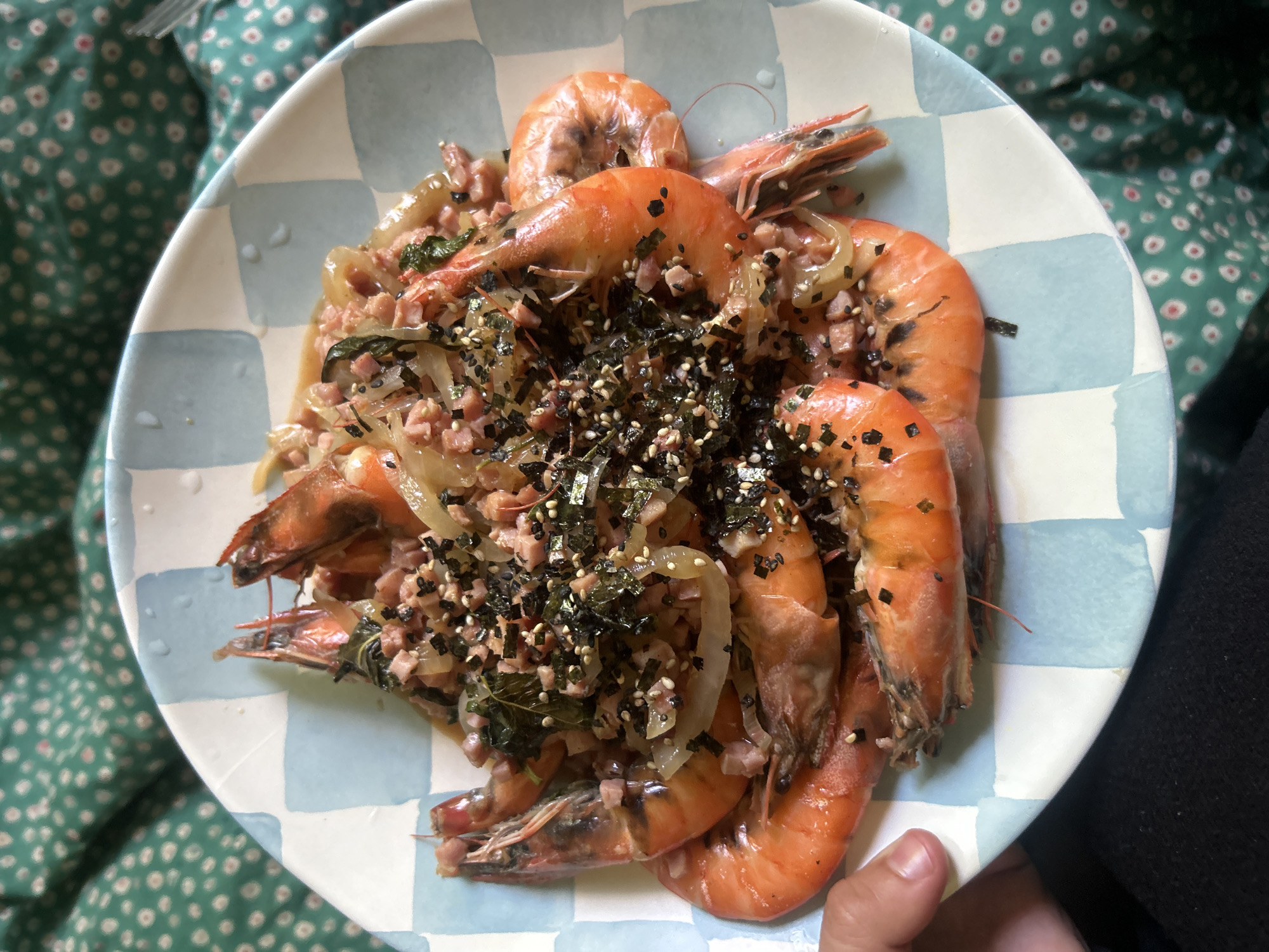 A plate of cooked shrimp is positioned atop a green floral print fabric, potentially a blanket or tablecloth. The shrimp is orange in color and topped with chopped ingredients, including seaweed and cubes of meat, possibly ham. Sliced onions are also visible among the shrimp. The plate itself features a blue and white checkered pattern. A hand is visible at the bottom right corner of the image. The setting appears to be indoors. There are no indications that this scene is specifically located in Los Angeles, nor are there people depicted beyond the hand holding the plate.