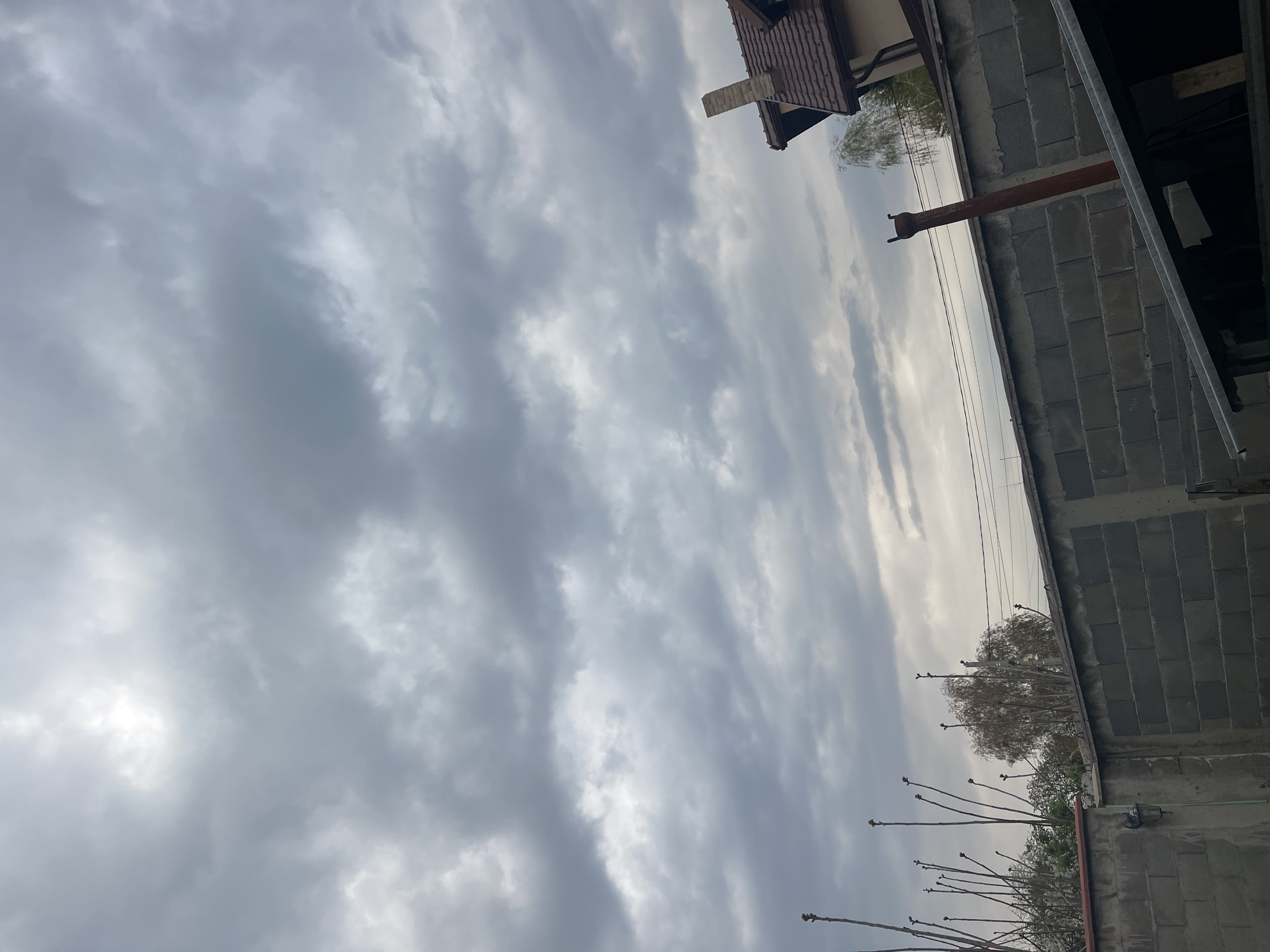 The image presents a low-angle, upward view of an overcast sky in what appears to be an urban or semi-urban setting, identified as Sofia, Bulgaria. The sky is largely covered by varying shades of grey clouds, with brighter, lighter patches visible, particularly towards the bottom-left, suggesting sunlight breaking through.
In the upper right portion of the frame, a building wall constructed from large, rectangular grey concrete blocks is prominently featured. A rustic, reddish-brown pipe or vent extends vertically from this wall into the cloudy sky. Adjacent to and slightly above this concrete structure, a section of a dark brown tiled roof, possibly belonging to another building, is visible, featuring a traditional style. Metal framing and supports are also seen along the upper edge of the concrete block wall.
Below the concrete wall and extending across the mid-right side of the image, several deciduous trees with mostly bare, vertical branches are present, suggesting a late winter or early spring season. Some younger branches display small buds or nascent foliage. Thin utility or electrical wires traverse horizontally across the mid-upper section of the cloudy sky. No people or specific activities are visible. The scene is static, capturing a cloudy daytime moment, likely portraying an area with ongoing construction or renovation, alongside established structures and natural elements. There is no visible text in the image.