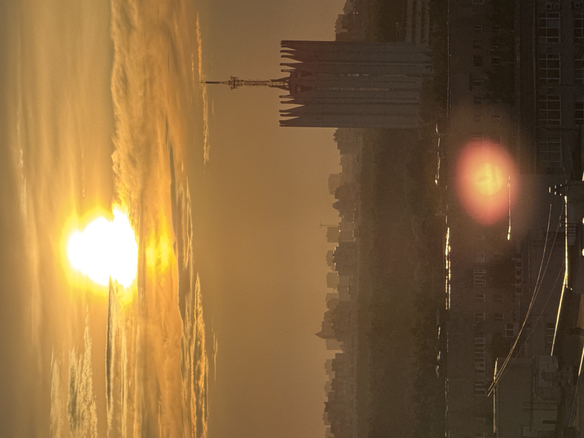The image displays an urban landscape at either sunrise or sunset, bathed in a warm golden-orange light. A brilliant, overexposed sun is visible low in the sky on the left side of the frame, its intense glow creating bright reflections and contributing to the overall warm tone. Horizontal streaks of clouds, illuminated by the sun, stretch across the sky, becoming denser around the sun itself.
In the mid-ground, a prominent, tall building with a distinctive, angular or finned top stands silhouetted against the bright sky. A slender communication tower or antenna extends vertically from its summit. Below and to the right of this central structure, a hazy silhouette of a sprawling city skyline stretches across the horizon.
The right side of the image features darker, less distinct foreground buildings, some with visible windows. A pronounced, reddish-orange circular lens flare is visible over these structures, suggesting light reflecting off the camera lens. Several utility lines or wires crisscross the upper right portion of the frame, catching glints of the golden light. No people are visible, and there is no discernible text. The scene captures a serene "golden hour" moment in an urban environment, consistent with the context of unknown Russia.