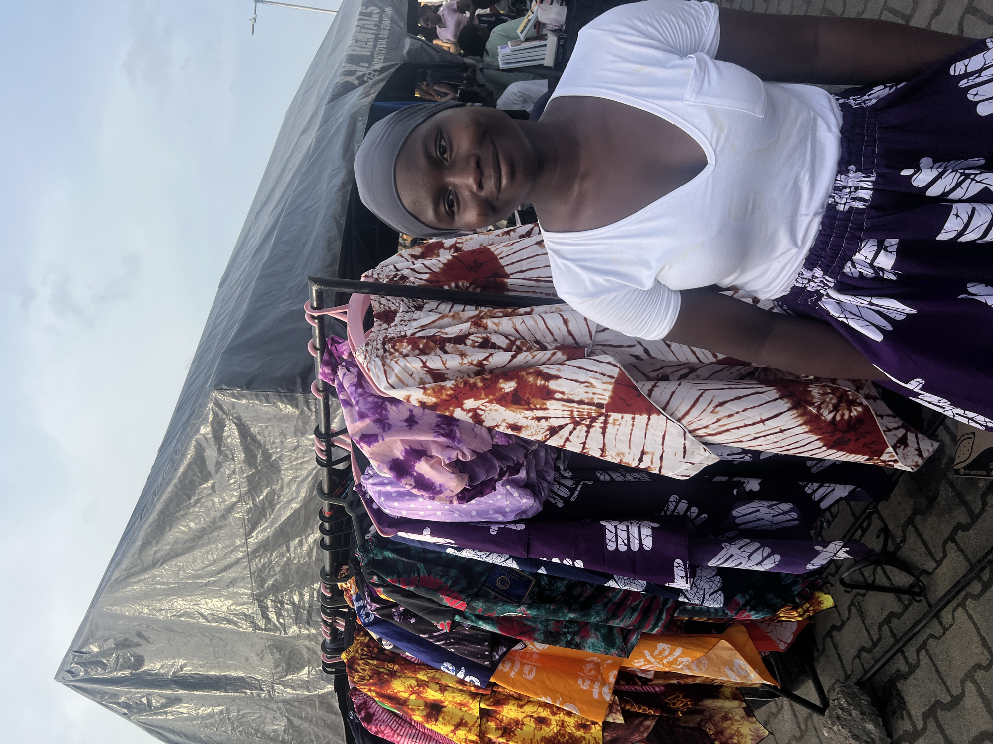 A woman wearing a white t-shirt and a purple and white patterned skirt stands behind a rack of colorful printed fabrics. The rack is filled with folded textiles of varying colors and patterns, hanging on black and pink hangers. The woman has a gray headscarf. A gray canopy provides shade over the area. The ground is paved with patterned gray bricks. In the background, blurred figures of people can be seen. The location appears to be an outdoor market or vendor area, likely in Ijanikin, Nigeria.