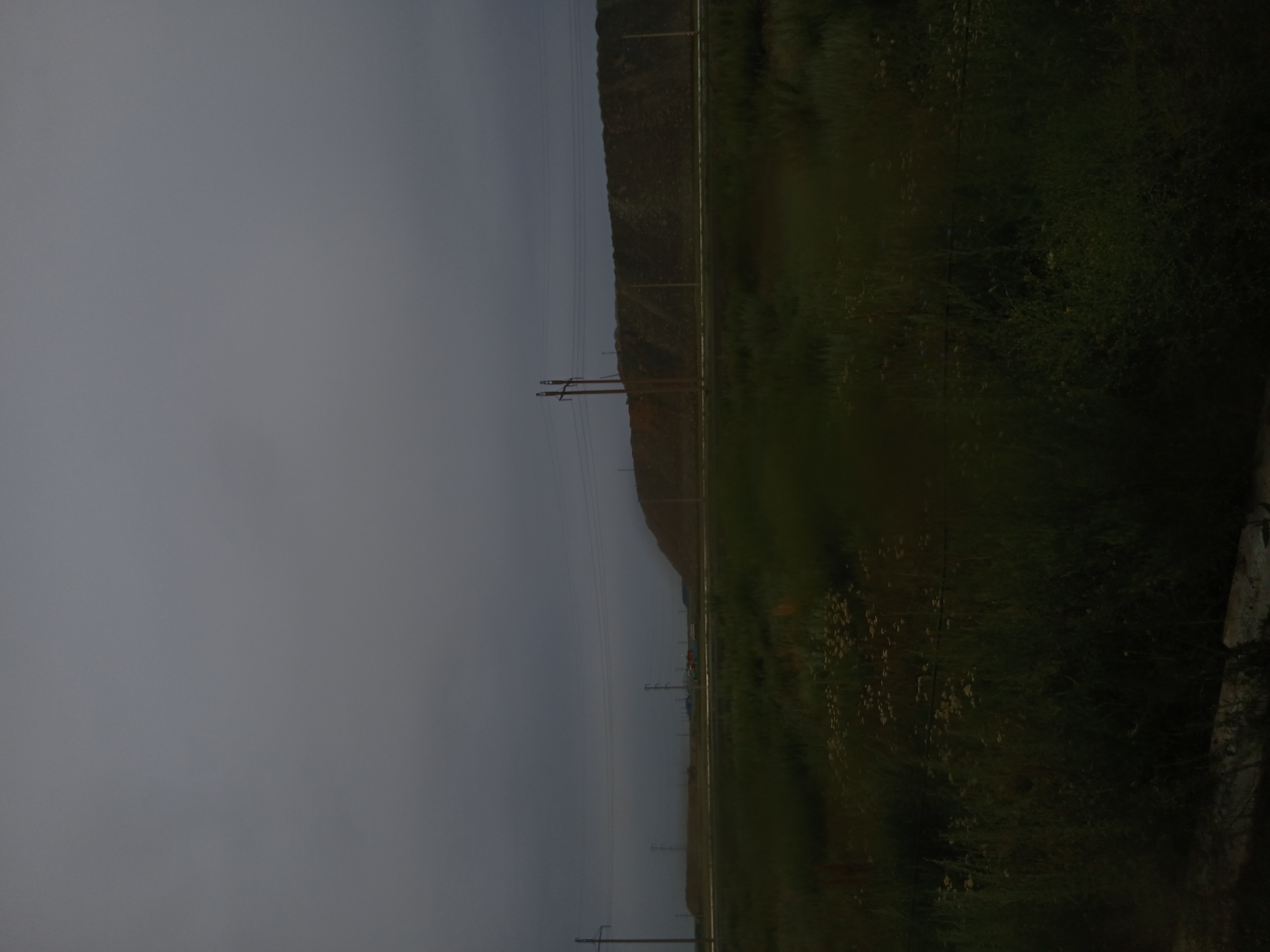 This image depicts an overcast, dimly lit scene in the city of Hromtau, Kazakhstan, likely on the outskirts or industrial zone. The foreground is dominated by tall, dark green grass with scattered small white wildflowers. A faint, horizontal line, possibly a fence or buried cable, is visible cutting through the vegetation.
In the middle ground, a flat, open area stretches towards a large, man-made hill or mound that occupies a significant portion of the background. This mound appears to be composed of earth or waste material, with visible textures and variations in color, ranging from dark brown to lighter ochre tones. Power lines and utility poles are scattered across the landscape, with one prominent wooden pole in the left middle ground and several others more distant. The sky is a uniform, dull gray, indicating heavy cloud cover and a lack of direct sunlight. The overall impression is one of a desolate, possibly industrial or post-industrial landscape under a somber sky.