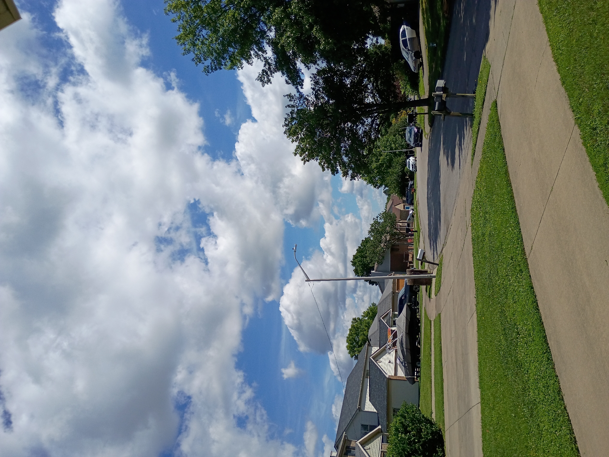 The image depicts a suburban street scene in Nicholasville, United States of America, under a partly cloudy sky. The foreground shows a concrete sidewalk and a well-maintained green lawn. Beyond the lawn, a paved residential street curves into the distance, lined with houses and trees. Several cars are parked on the street and in driveways. One house features a boat covered with a gray tarp on a trailer in its driveway. A lamppost stands near the intersection, and a cluster of mailboxes is visible on the right side of the street. The environment appears to be a typical residential neighborhood on a sunny day.
