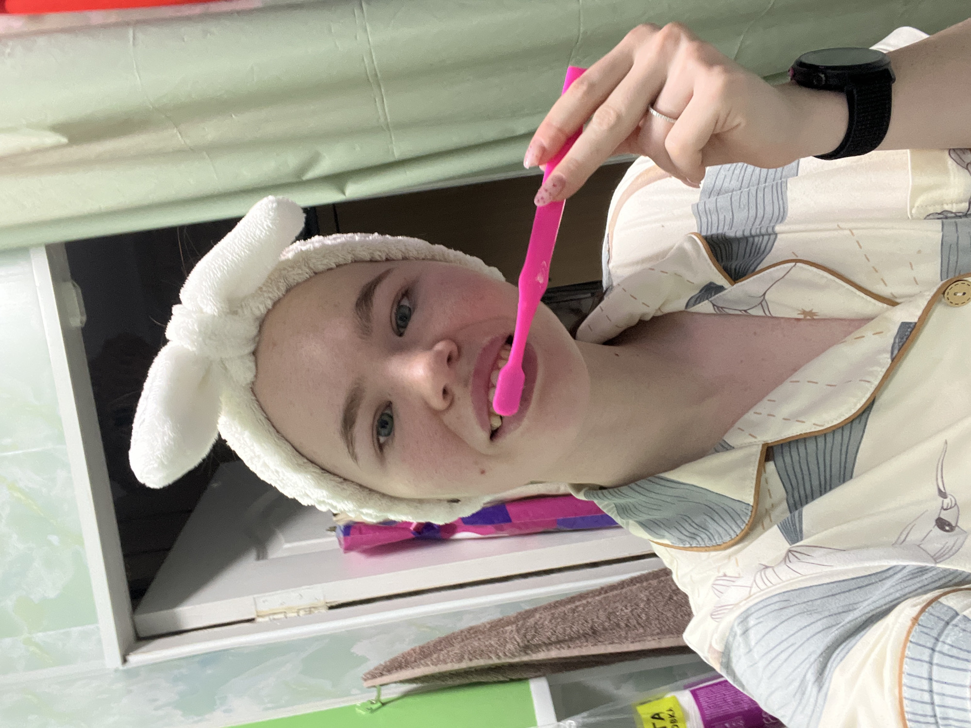 A young person is captured in a close-up, casual self-portrait indoors, engaged in a personal hygiene routine. The subject, likely female, has fair skin, light-colored eyes, and light hair mostly covered by a white, fluffy bunny-ear headband.
They are holding a bright pink toothbrush to their mouth with their right hand, mid-action of brushing their teeth. A subtle design or glitter is visible on their fingernails. On their left hand, a silver ring adorns their ring finger, and a dark-banded smartwatch is worn on their wrist.
The person is dressed in patterned pajamas, featuring a light background with blue-grey stripes and abstract designs. The setting appears to be a bathroom or bedroom, indicated by the activity and background elements. Behind the person, a light-colored blind or curtain hangs, and white cabinetry or shelving is visible. A brown textured towel and various other items, including a pink and purple package, are present on a surface in the background.
The image conveys a moment of routine, possibly in the morning or evening. The person displays a mild, pleasant expression. Partially visible Cyrillic text, reading "ТА ОДКА", can be seen on a package in the lower right, consistent with the location context of Chaikovsky, Russia.