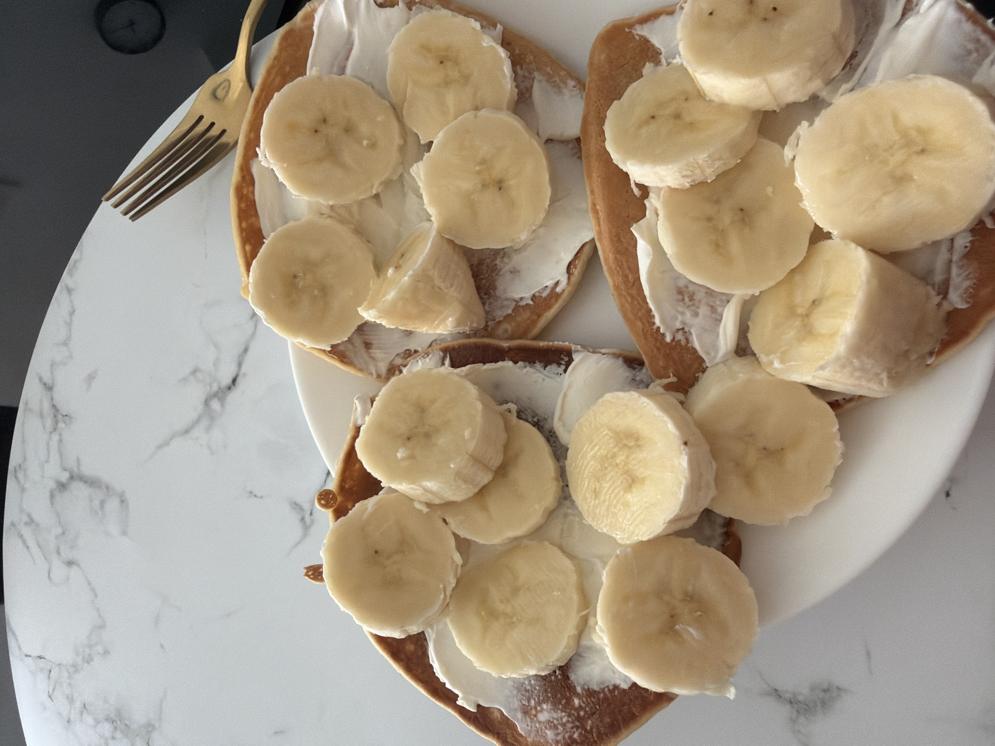 A close-up, high-angle shot captures three banana and cream cheese crepes arranged on a white plate. The crepes are golden brown and appear to be slightly folded or overlapping. Each crepe is generously spread with a white cream cheese frosting, and topped with numerous round slices of fresh banana. A gold-colored fork rests on the right side of the frame, positioned above the crepes, with its tines pointing towards the food. The background consists of a white marble-patterned surface. The scene is set on a table in Finestrat, Spain.