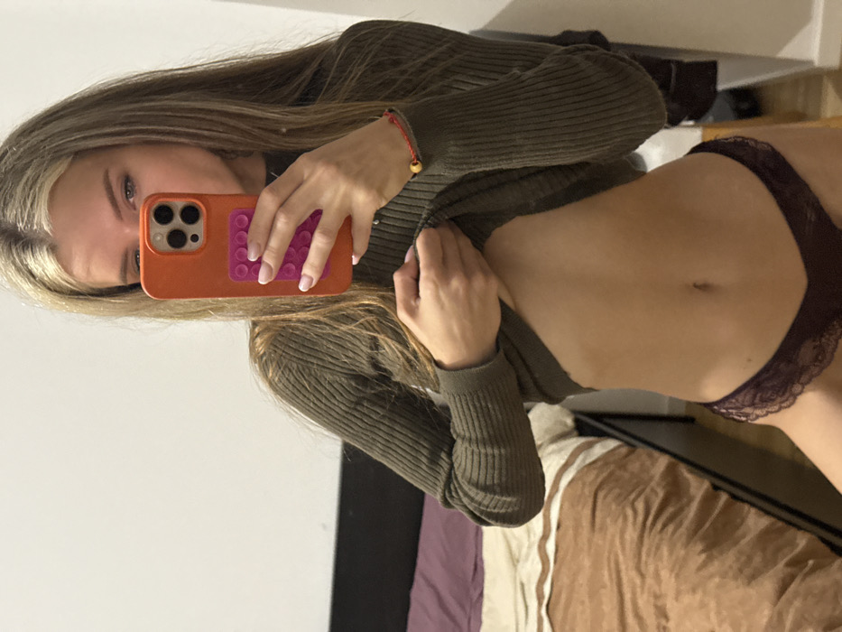 A woman takes a selfie in a mirror, holding an orange iPhone with a pink pop-socket. She wears a fitted, green long-sleeved shirt and dark purple, lacy underwear. The image captures her from the chest up, angled slightly to the left. A red string bracelet is visible on her left wrist. The background includes a bed with brown and purple bedding and a light-colored wall.