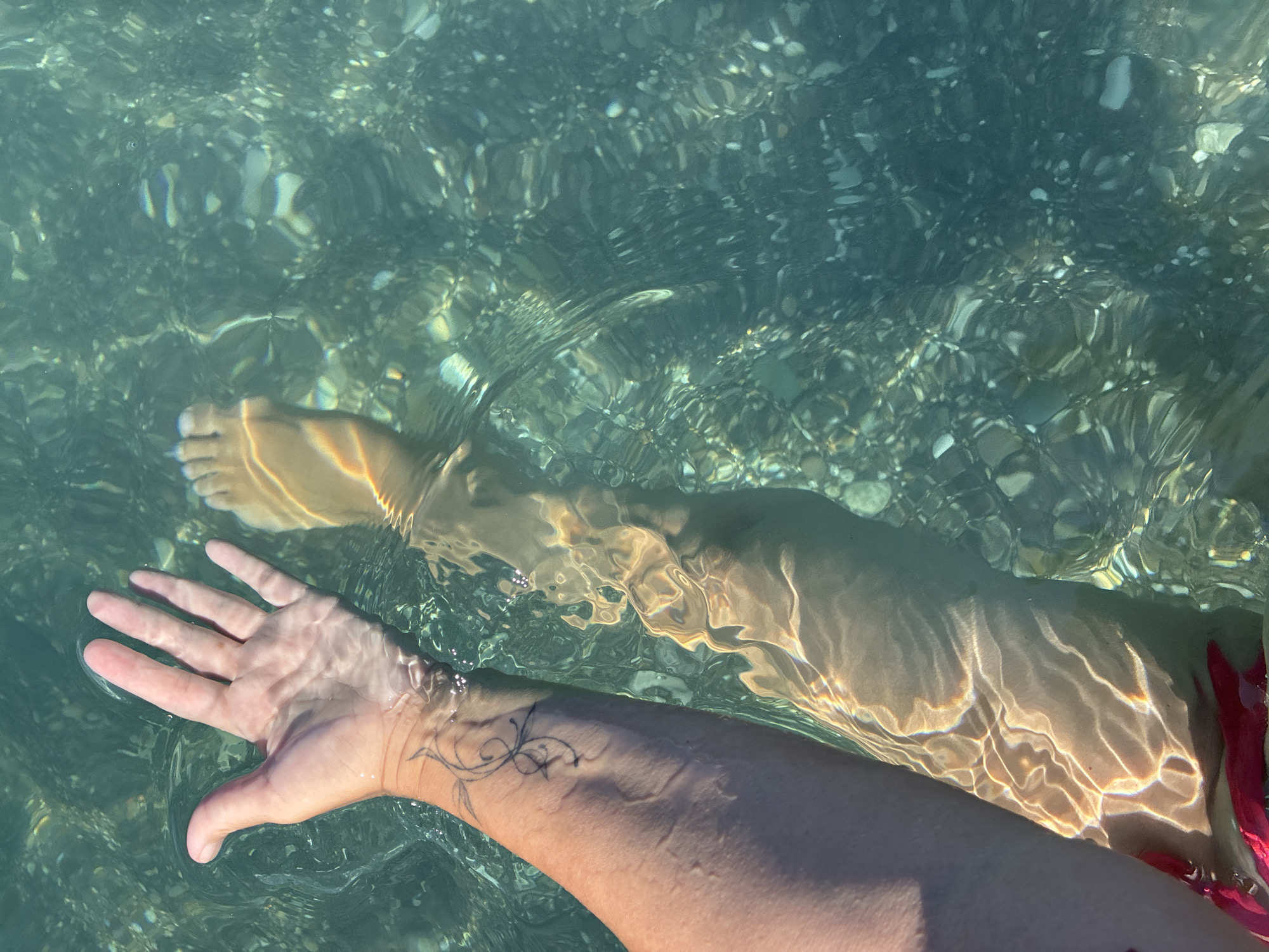 The image shows a person's arm and leg submerged in clear, shallow water. The water is turquoise and contains small, smooth pebbles on the seabed, illuminated by sunlight that creates dappled patterns on the skin. The person has a small tattoo on their left forearm. The person's right foot is also visible, with toes extending into the frame. The overall impression is of a relaxed, summery scene. This image is not specific to Sochi, Russia.