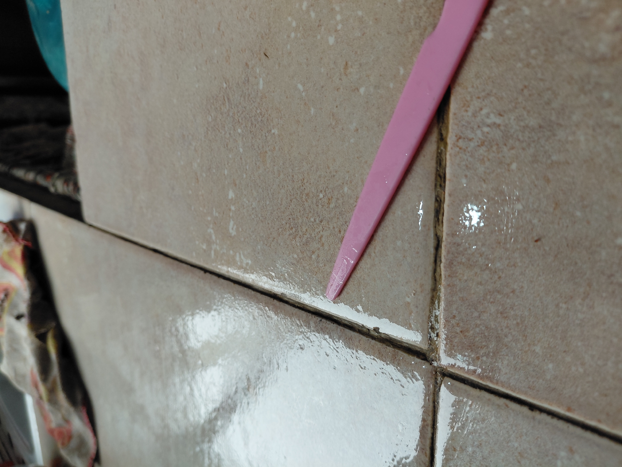 The image captures a close-up view of a tiled surface undergoing cleaning or renovation, likely in a kitchen or bathroom in Santa Ana, Venezuela. The main subject is a section of light brown or beige tiles, some of which appear wet and reflective. A prominent pink, pointed tool, resembling a scraper or a grout tool, is positioned within a grout line.
The activity depicted is the cleaning or application of new grout. One of the horizontal grout lines shows a fresh, white substance, possibly new grout or a cleaning paste, being applied or spread by the pink tool. Other grout lines are visibly darker and appear dirty or discolored, suggesting mold or grime buildup, contrasting with the cleaned or newly treated section. The tiles themselves have a slightly textured, granular appearance with numerous small white specks scattered across their surface. The wetness on the tiles and within the grout lines indicates that water or a cleaning solution is being used.
In the blurred background on the left, hints of a blue object and some fabric or a towel are visible, further suggesting a domestic cleaning scenario. The focus is tightly on the tiles and the tool, emphasizing the cleaning process. There is no visible text in the image. The scene conveys a sense of diligent upkeep or a home improvement task.