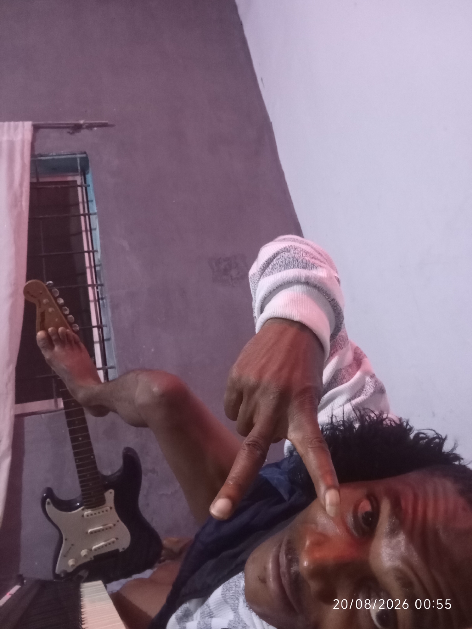 In a dimly lit room, a person lies on their back with their left leg extended and bent at the knee. A black electric guitar rests between their legs, with the headstock positioned near their foot. The person is wearing a striped sweater and appears to be making a peace sign with their right hand, which is held near their face. The background shows a wall with a window covered by a grate and a curtain. A timestamp in the bottom right corner reads "20/08/2026 00:55". The scene appears to be indoors, likely a residential setting. The specific location of Adankolo, Nigeria, is not discernible from the visual elements of the image itself.