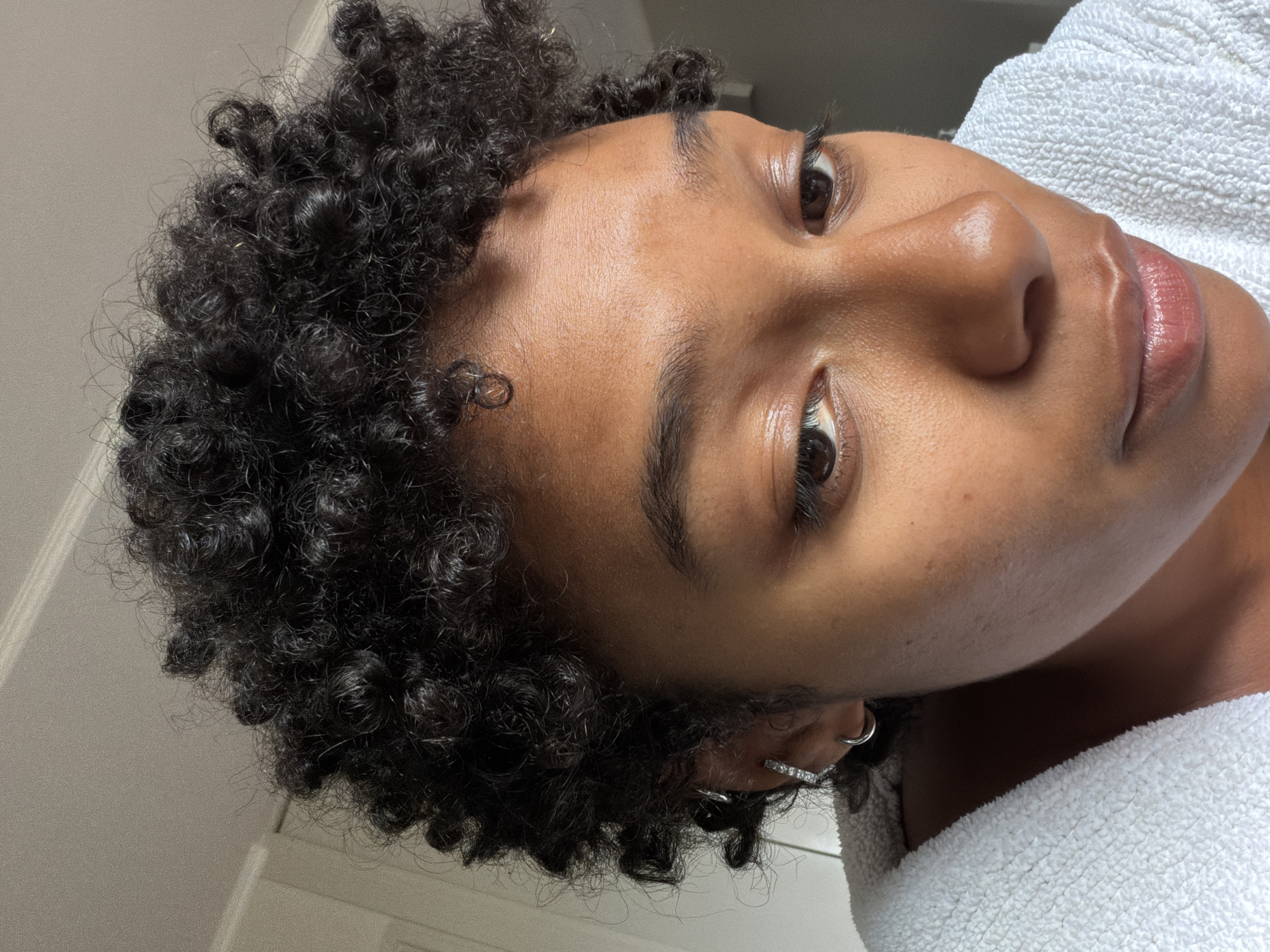 A close-up, rotated portrait shot captures a person with dark, tightly coiled curly hair and a medium-dark complexion, likely taken indoors in Champs-sur-Marne, France. The subject's head is tilted, and they are looking upwards, with their gaze directed slightly away from the camera. Their dark brown eyes are open, and their expression appears calm and contemplative.
The focus is on their face, showcasing dewy, hydrated skin that reflects light, particularly on the forehead, nose bridge, and cheekbones. Their lips have a natural pinkish hue and a glossy finish. Dark eyebrows are well-defined. On the visible ear, multiple silver earrings are present, including a simple hoop and a decorative ear cuff with small, clear stones.
The person is wearing what appears to be a soft, white terry cloth robe or towel, visible on their right shoulder and neck area, suggesting a relaxed or post-bath/shower setting. The background is uniformly light gray or off-white, providing a clean, uncluttered backdrop that keeps attention on the subject. The lighting is soft and even, highlighting the textures of the hair and the sheen on the skin without harsh shadows. No text is visible in the image.