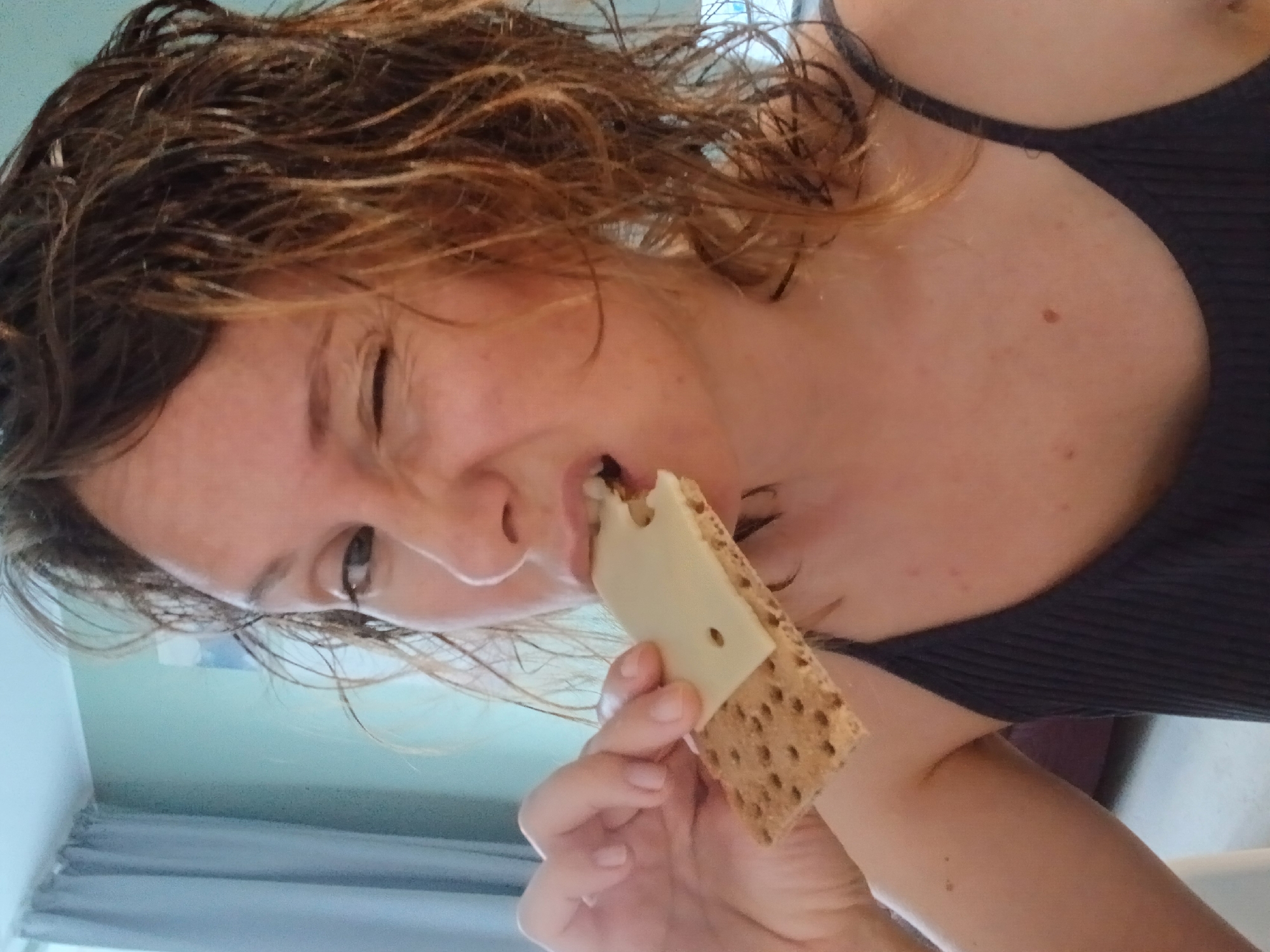 A close-up shot captures a woman with messy, light brown hair biting into a cracker topped with a slice of cheese. She is winking, with her mouth slightly open, and her left hand is holding the cracker. She is wearing a dark, ribbed tank top. The background is blurred, showing a teal wall and a grey curtain. The scene appears to be indoors, possibly in an apartment. This image was taken in the city of Yekaterinburg, Russia.