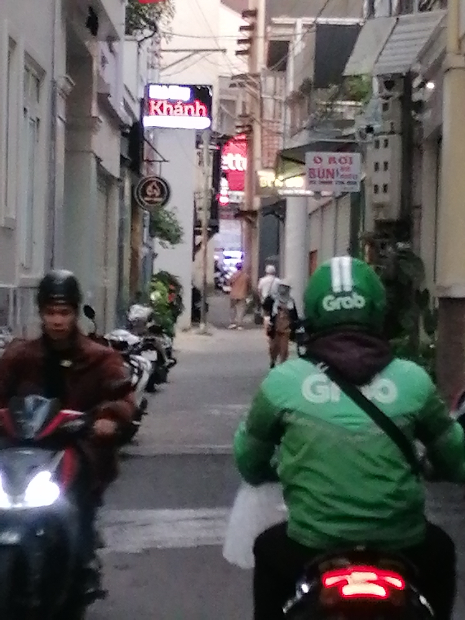 The image depicts a street scene in Da Lat, Vietnam, likely during daylight hours. In the foreground, a person wearing a green "Grab" branded jacket and helmet is riding a motorcycle. The Grab driver is the central subject, with their back to the camera. To the left, another person on a motorcycle, wearing a dark helmet and a reddish-brown jacket, is also visible, facing forward. The street is lined with buildings, some of which have signs, including one with "Khánh" in neon letters. Several motorcycles are parked or in motion on the street, indicating a common mode of transportation. The environment appears to be an urban setting with a narrow street and pedestrian activity in the background. The overall impression is of a bustling city street.