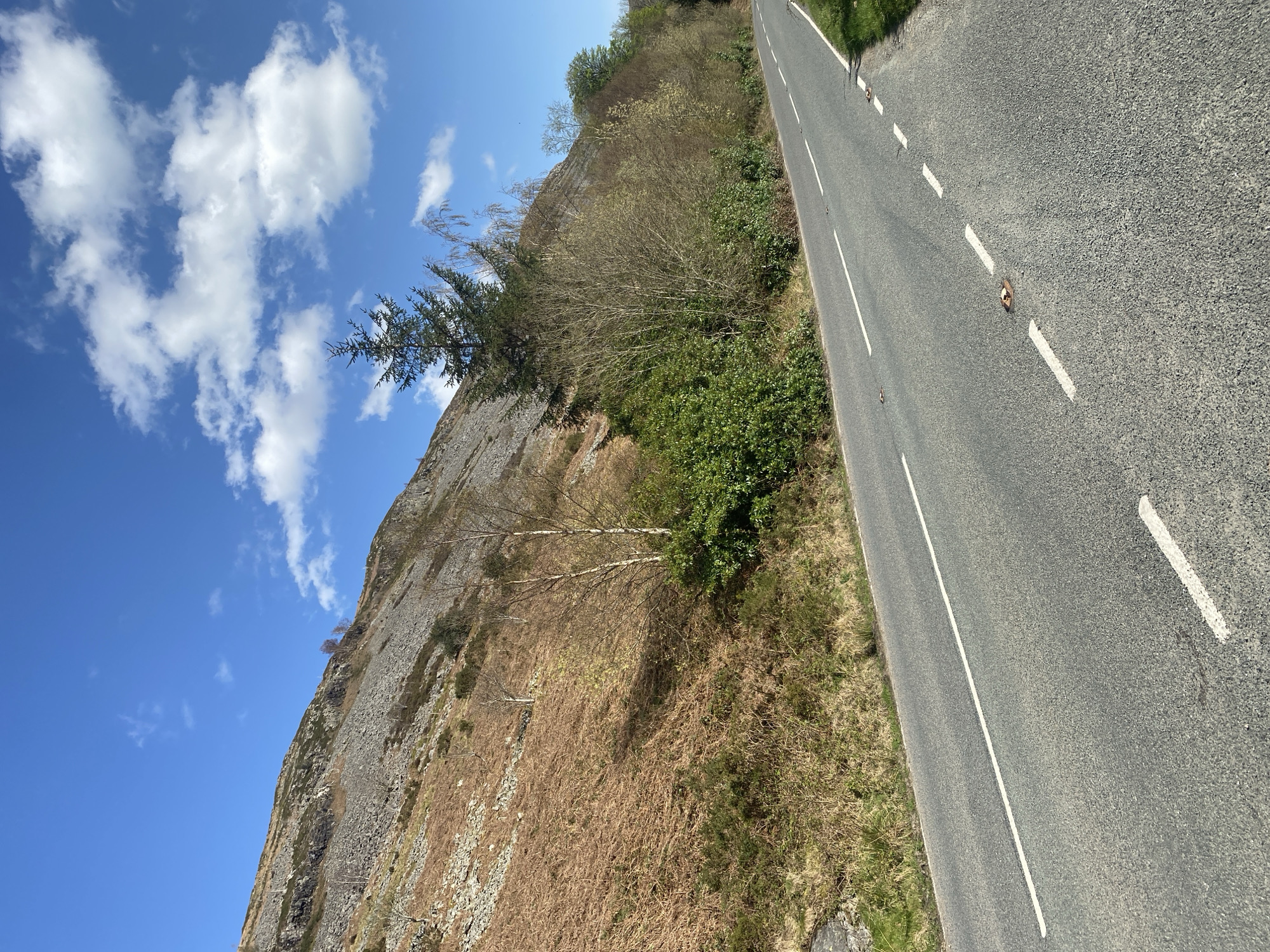 The image depicts a road running alongside a steep, rocky hillside in Llangynog, United Kingdom. The road is paved with gray asphalt and has white dashed lane markings and cat's eye reflectors. The hillside is covered in brown and green vegetation, with rocky outcrops visible. A few trees are scattered across the hillside. The sky is blue with scattered white clouds. There are no people visible in the image.