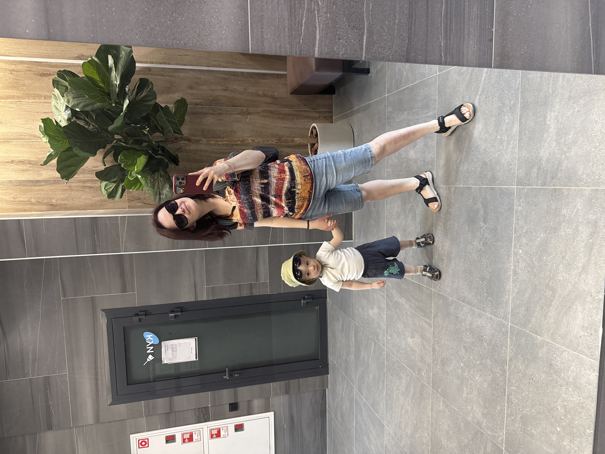 A woman and a child are reflected in a wall-sized mirror in an interior space with gray tiles. The woman wears sunglasses, a patterned top, and denim shorts, and holds a smartphone. The child wears a bucket hat, sunglasses, a t-shirt, and shorts with a dinosaur patch. A potted plant is visible in the upper left of the reflection. A dark gray door and a white fire safety box are against the wall next to the mirror. A bench is slightly visible at the top of the mirror. The location may be Izhevsk, Russia, but this cannot be confirmed from the image.