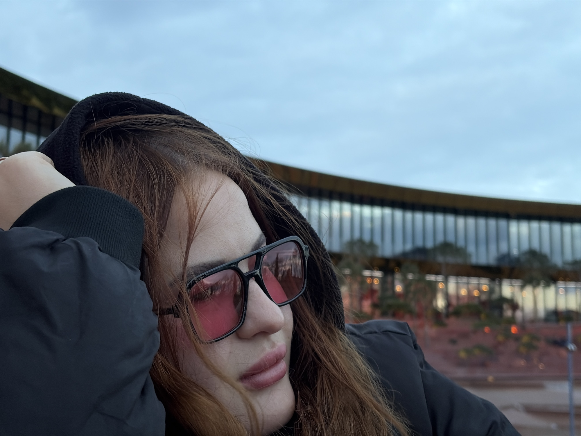 The image captures a person, likely female, in a close-up, angled shot from their right side. The subject wears a dark, possibly black, hooded jacket, with long, reddish-brown hair visible around their face and under the hood. They are wearing large, black-framed sunglasses with prominent pink-tinted lenses, through which blurred reflections are visible. The person's lips appear full and softly colored. Their right arm is raised, with the hand positioned near their head, possibly adjusting the hood or resting.
The background features an overcast, light blue-grey sky dominating the upper portion of the frame. Below the sky, a modern architectural structure with a curved facade is partially visible. This building has multiple windows or glass panels and a distinct horizontal golden-brown band across its upper section. Below the building, out-of-focus elements suggest a landscape with subtle lighting and blurry, warm-toned artificial lights, indicating either dusk, late afternoon, or an overall cloudy day. There is no visible text in the image.