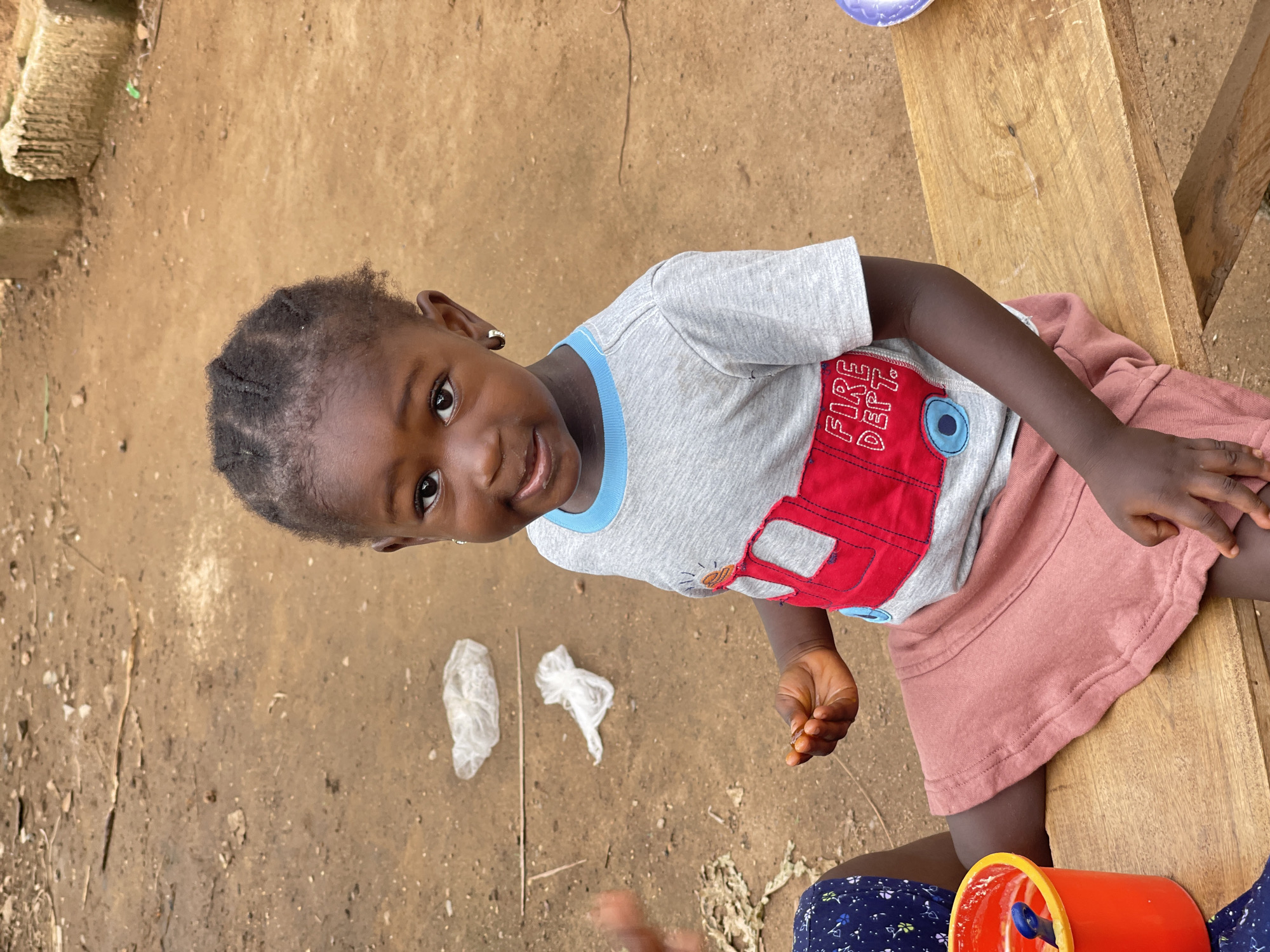A high-angle, portrait shot captures a young child sitting on a wooden bench against a dirt ground. The child has dark hair styled in braids, dark eyes, and brown skin. She is wearing a gray t-shirt with blue trim and a red fire engine graphic, as well as a pink skirt. The child is smiling and looking directly at the camera. In the background, there is dirt ground, as well as plastic trash and what appears to be a red pail. The location is Akwanga, Nigeria.