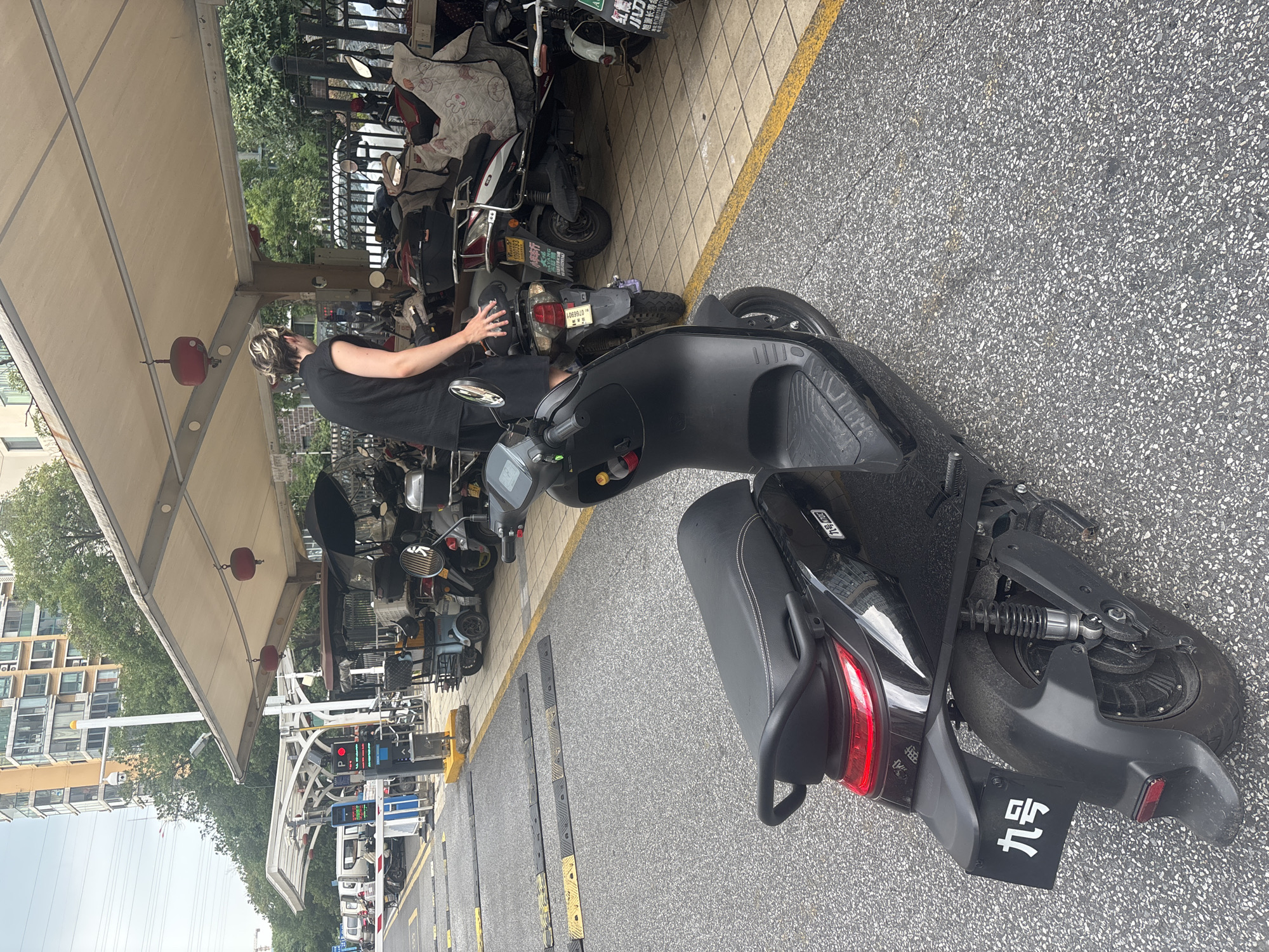This image depicts an outdoor electric scooter parking area located in Suzhou Industrial Park, China. A person with short, two-toned hair and wearing a black sleeveless top and shorts stands next to a black electric scooter. They are reaching towards the rear of a parked scooter. In the background, a row of various electric scooters and mopeds are parked under a covered shelter. The ground is asphalt with yellow and black speed bumps visible. Beyond the shelter, buildings and trees are present. A security camera is mounted on a pole in the far background.