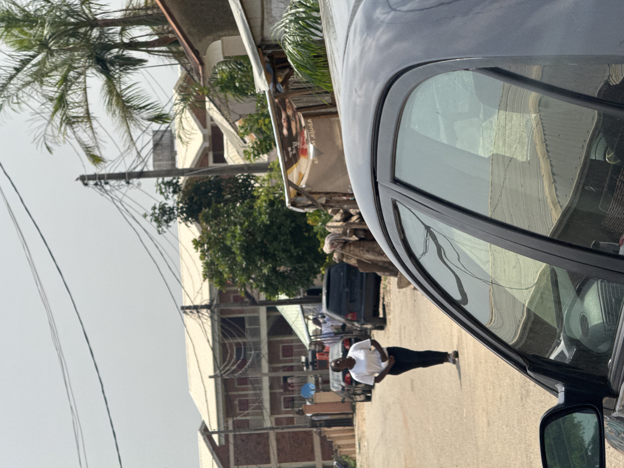 A person wearing a white t-shirt and black pants walks down a street in a town, possibly Abuja, Nigeria. The person is in the foreground and is walking away from the camera. The street is paved with asphalt and lined with buildings on either side. The buildings appear to be residential and commercial, with some of them having balconies and multiple stories. There are also some trees and power lines visible in the background. A dark-colored car is parked on the side of the road, and its reflection can be seen in the window of another car, which is partially in the frame on the right side. The weather appears to be clear and sunny, suggesting it's daytime. The scene is typical of a moderately developed urban or suburban area. There is no visible text in the image.