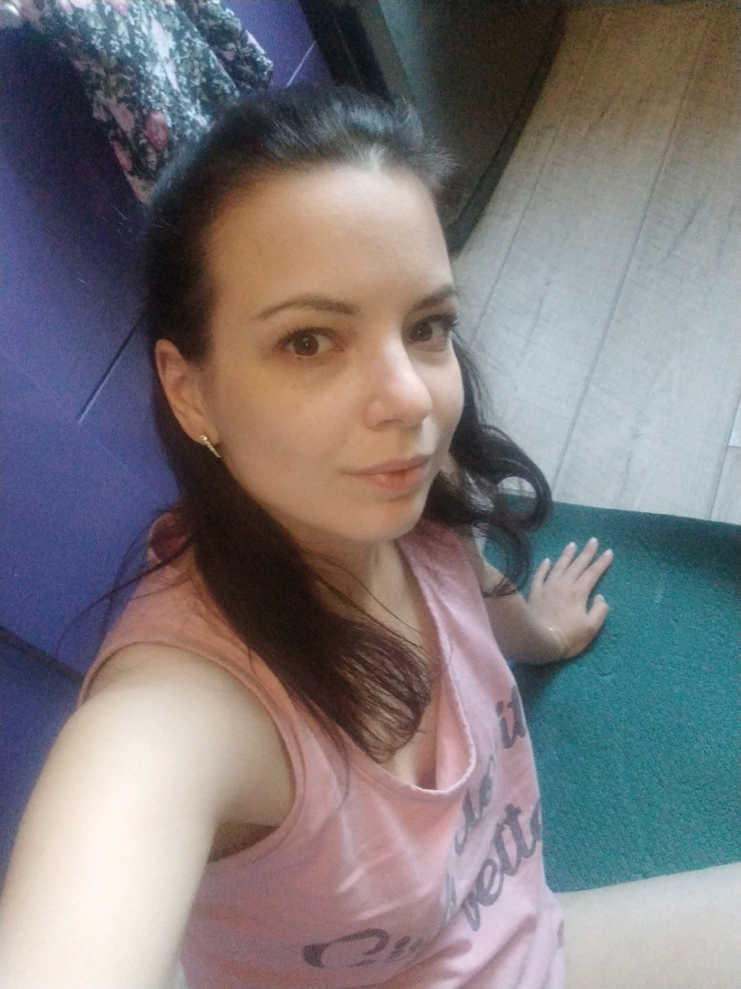 A young woman with dark hair and brown eyes is sitting indoors, looking at the camera. She is wearing a pink tank top with a graphic print. Her right hand is resting on a turquoise mat. The background shows a wooden floor and a purple surface.