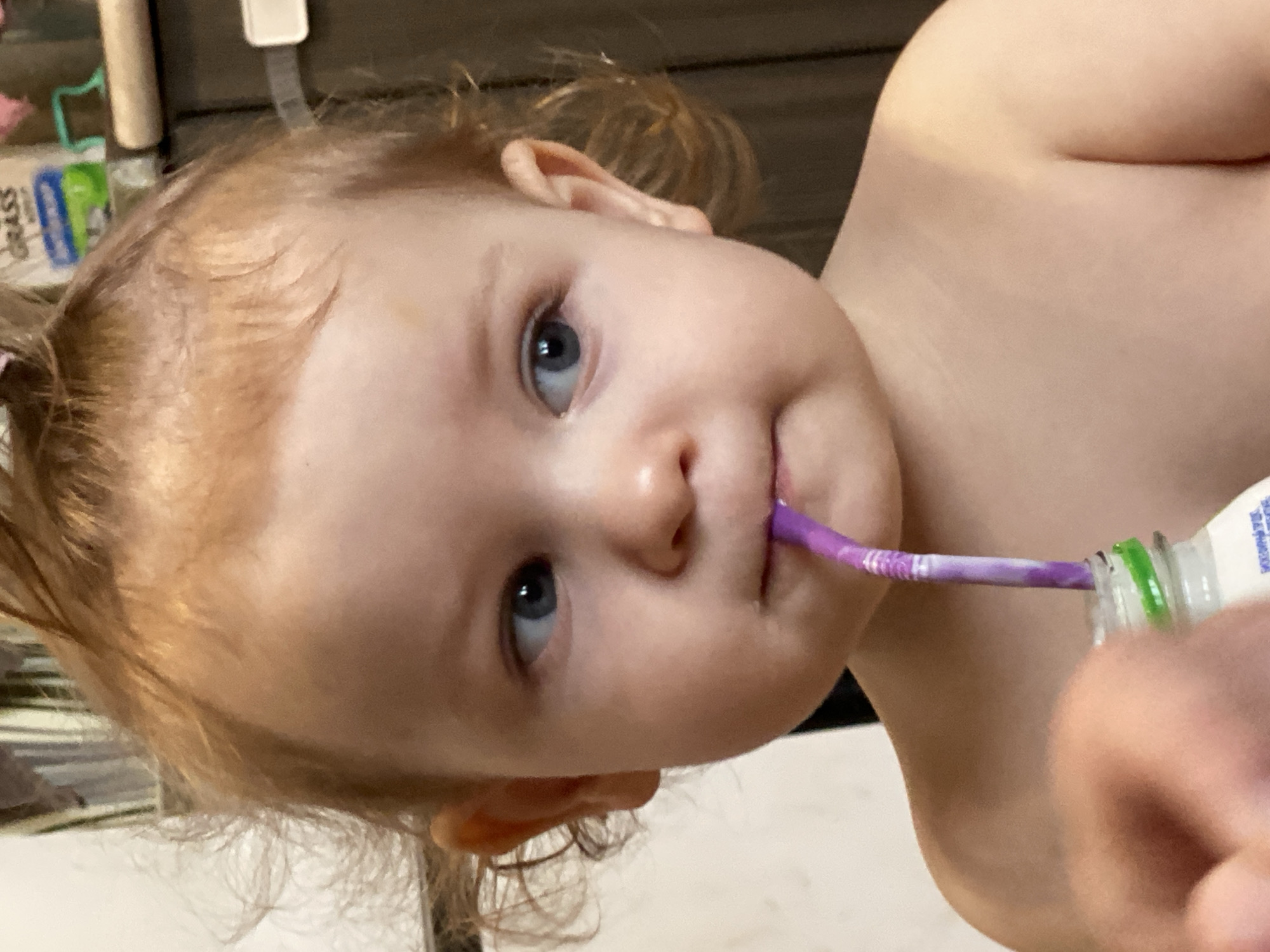 A close-up shot shows a light-skinned toddler with light brown hair, likely indoors. The child has blue eyes and is holding a purple and white toothbrush in their mouth while seemingly consuming toothpaste directly from a bottle. The background is slightly out of focus but suggests a domestic interior.