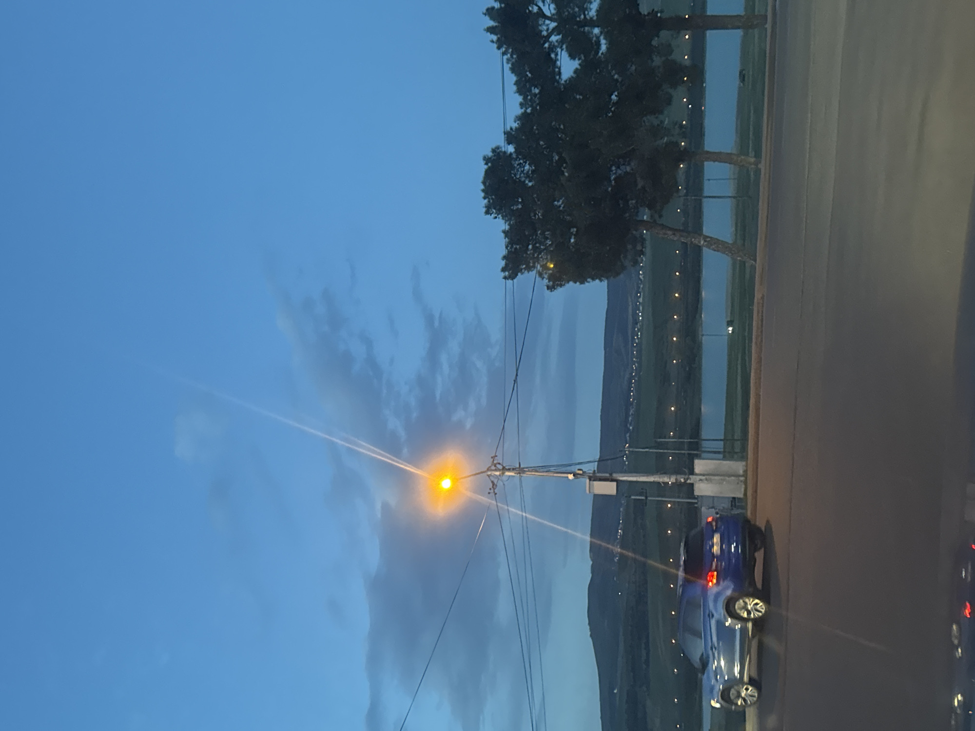 The image captures an outdoor twilight or early night scene, likely in a suburban or semi-rural area, consistent with the provided context of Tbilisi, Georgia. The sky, a deep blue transitioning to lighter hues, fills the upper half of the frame, adorned with scattered, wispy clouds. A prominent street light, affixed to a utility pole on the right side of the image, emits a bright yellow-orange glow with noticeable light rays, indicating the low light conditions. Electrical wires extend from the pole, crossing the mid-ground.
Below the utility pole, a dark blue car is parked on the edge of an asphalt road that runs along the lower right portion of the frame. Its rear lights are faintly visible. To the left of the road, a tranquil body of water, possibly a lake or river, stretches into the distance, reflecting the twilight sky. Along the far bank of the water, a range of rolling hills or mountains forms the horizon, silhouetted against the lighter sky. Numerous small, incandescent lights are visible twinkling across the distant landscape, suggesting scattered buildings or other artificial lighting along the shore or on the hills. Several dark, leafy trees stand along the roadside and by the water's edge, their forms largely silhouetted. No people are visible, and no specific activity is taking place. There is no discernible text within the image.