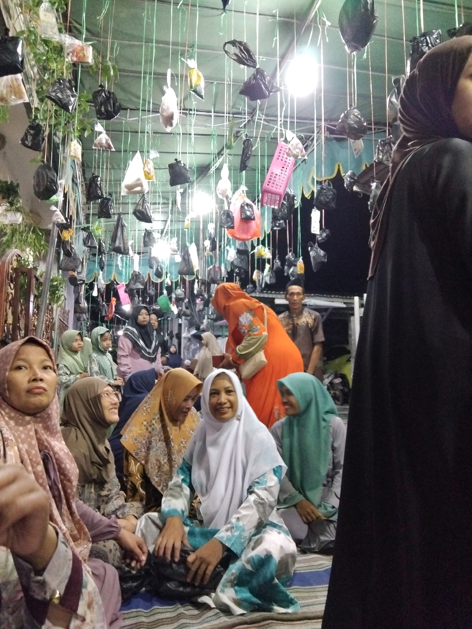 The image depicts an outdoor gathering at night in Kepuh Kiriman, Indonesia, possibly a celebration or event. The scene is characterized by a dense canopy of items hanging from overhead strings, including black plastic bags, white plastic bags, and some pink and orange items. These hangings create a cluttered, festive, or perhaps symbolic atmosphere.
In the foreground and mid-ground, a group of people, predominantly women, are seated on the ground. They are all wearing headscarves (hijabs) in various colors and patterns, including pink, light green, brown, yellow with intricate patterns, and white. Their clothing also varies, with some wearing patterned dresses and others solid colors. Several individuals are looking towards the camera or interacting with each other. One woman in the front left is wearing a pink patterned hijab and a matching patterned dress, her face is turned towards the camera. Next to her, a woman in a brown hijab and patterned dress is smiling. In the center, a woman in a white hijab and a green and white patterned dress is seated. To her right, a woman in a green hijab is also seated.
Further back, a man in a brown patterned shirt is visible, looking towards the left of the frame. An orange garment is worn by a woman partially obscured by the foreground subjects. The background reveals more of the hanging decorations and appears to be a dark, possibly unlit area, suggesting it is nighttime. The ground is covered with mats or rugs. The overall impression is a communal gathering taking place outdoors under a decorated ceiling of suspended items.
