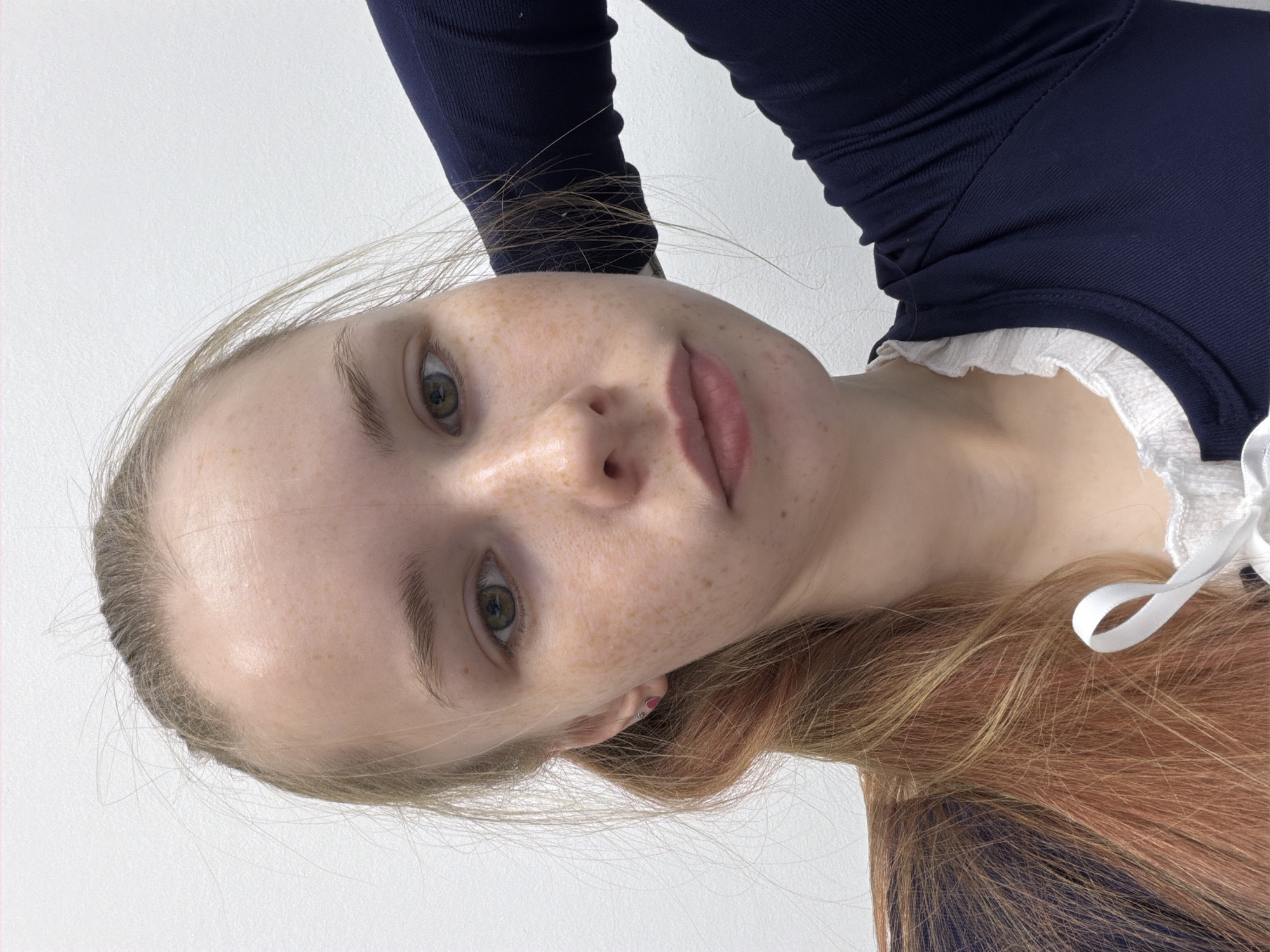A close-up, angled headshot depicts a fair-skinned person with light brown hair, freckles, and light hazel eyes. The person is wearing a dark blue garment with a white ruffled collar. Part of the person's hair is dyed a reddish-brown color. A small pink earring is visible in the person's left ear. The background is a plain white surface. The location in the city of Berezniki, Russia, is not discernible from the image.