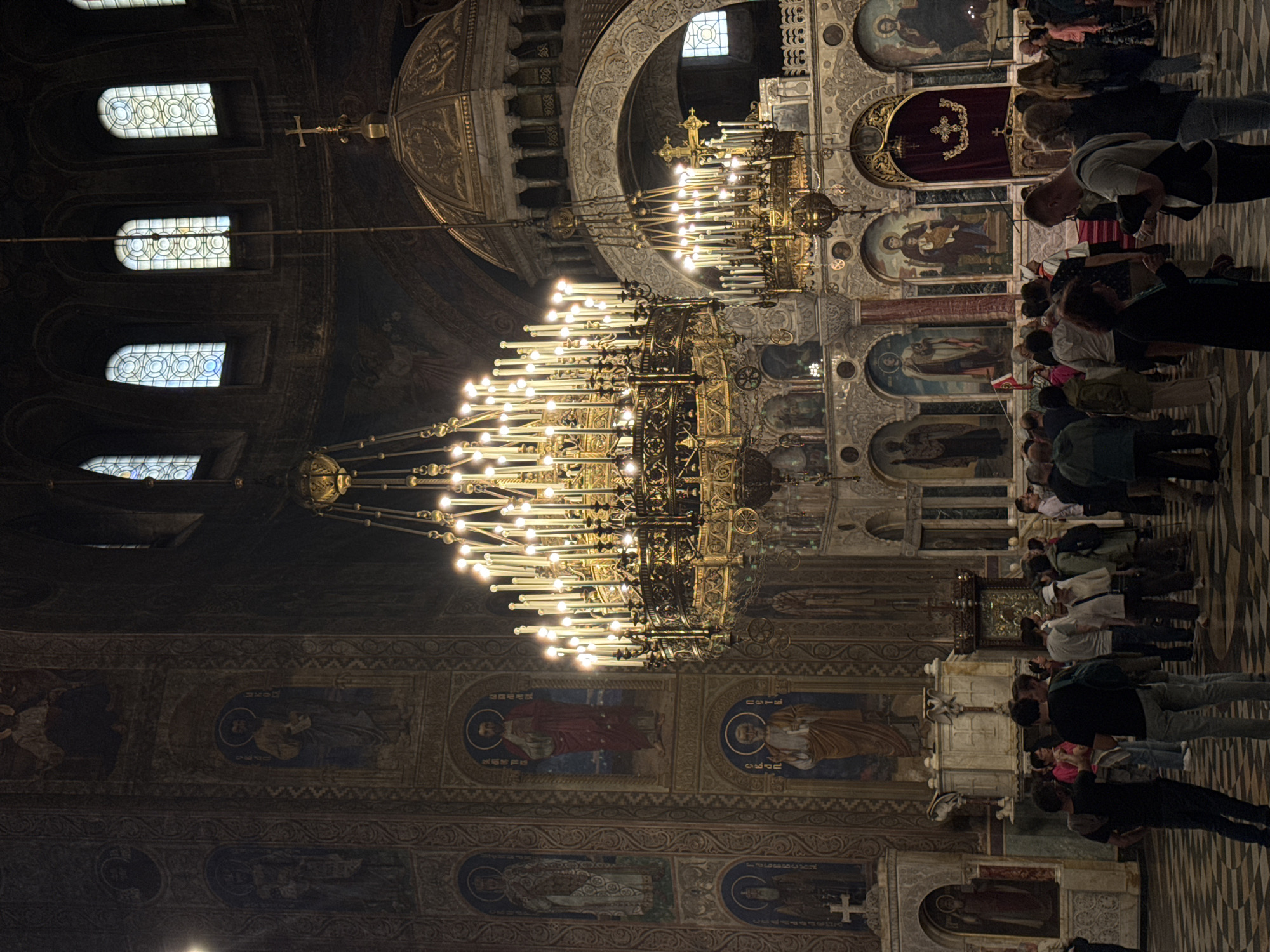 Here is a detailed description of the image:
The image captures the interior of an ornate church, likely in the city of Bucharest, Romania. Dominating the scene are two large, elaborate chandeliers. The larger one hangs prominently in the center, adorned with numerous lights.
The background of the image showcases the church's architecture, including arched windows at the top and intricate artwork, potentially mosaics or painted scenes, lining the walls. The paintings seem to depict religious figures.
A group of people is visible, observing the interior of the church. Some are looking up, possibly at the chandeliers or the artwork on the ceiling. Others seem to be engaged in conversation.
The image suggests a moment of quiet observation and reverence within a richly decorated and historically significant space. The presence of the chandeliers, artwork, and people all contribute to the atmosphere of the church.