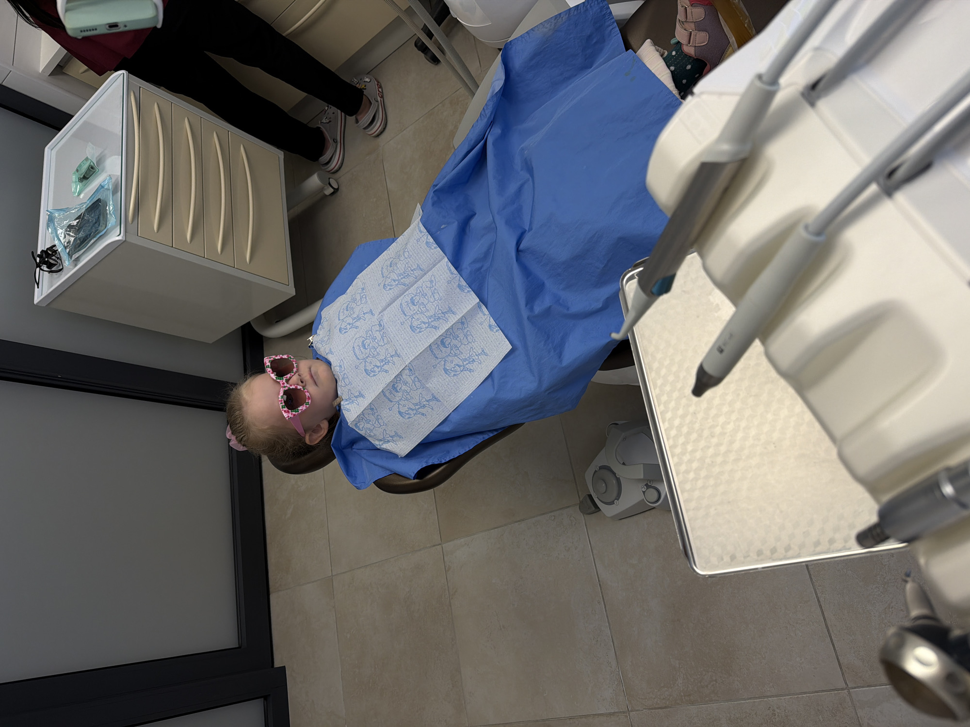 A young child with pink heart-shaped sunglasses lies in a dental chair covered with a blue paper drape and a paper bib with cartoon images. A dental hygienist or assistant stands nearby. The room is a dental office with beige tile flooring, a cabinet, dental instruments, and a dental chair. The location is assumed, but unconfirmed, to be in the city of Kamyshin, Russia.