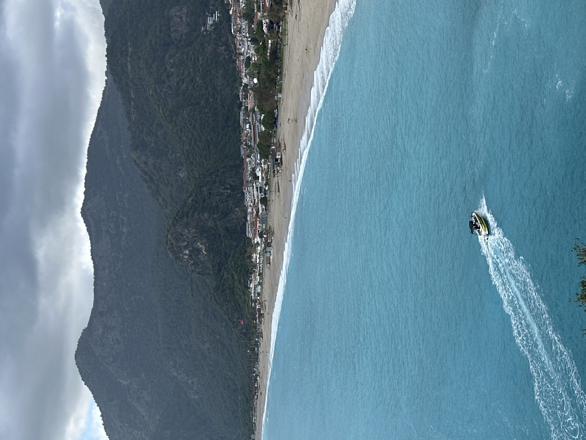 The image shows an aerial view of Ölüdeniz beach in Fethiye, Turkey. A beach runs along the center of the image, and turquoise water fills the right side. The shoreline is lined with buildings. To the left of the shoreline, a large mountain is visible. A jet ski is in the water, leaving a trail of white water behind it. The sky is partly cloudy.