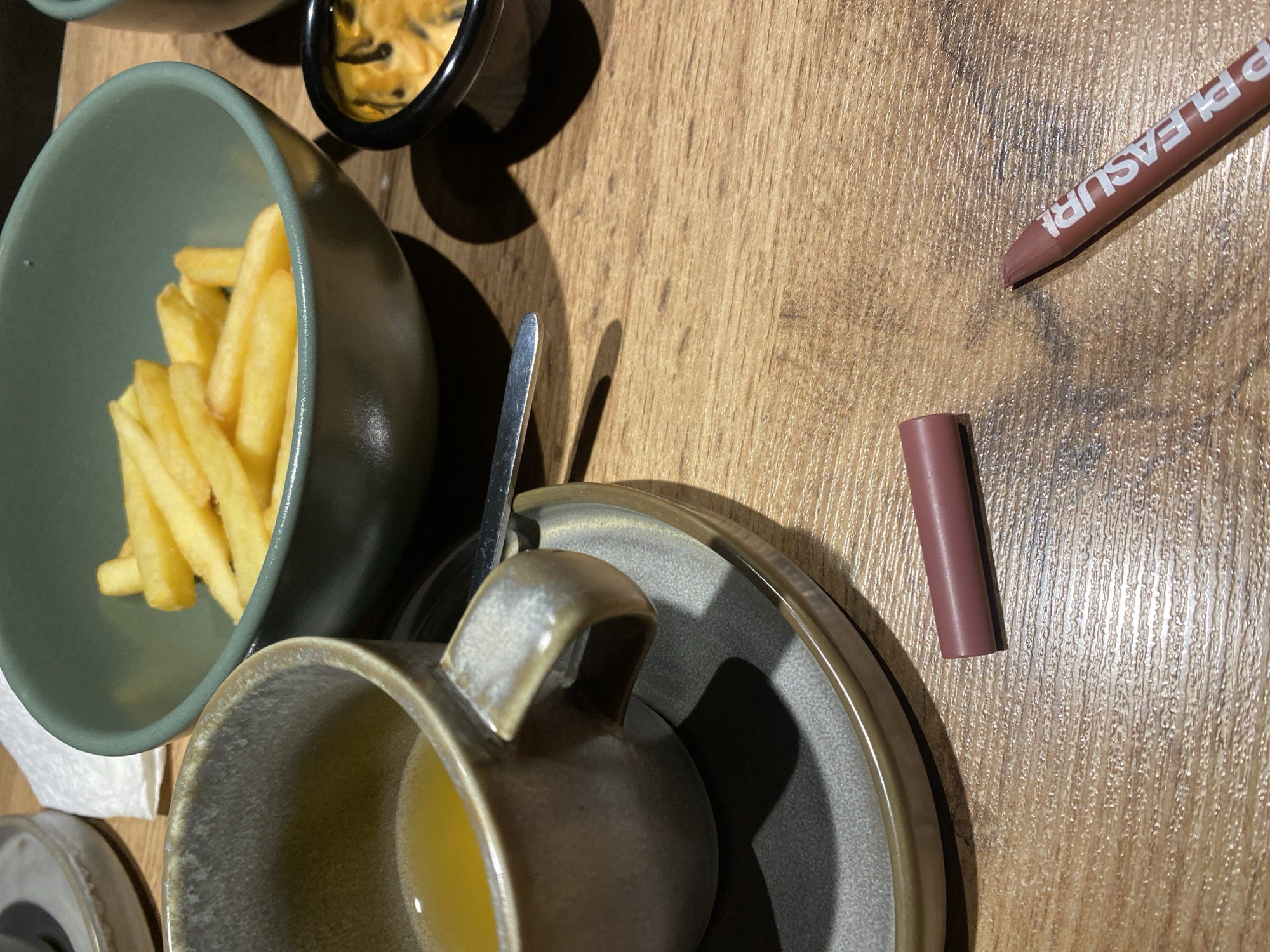 The image shows a table setting with a bowl of french fries, a small dish with dip, a cup with liquid, a spoon, and two makeup pencils on a wooden surface. The makeup pencils are brown, with one labeled "PLEASURE". The scene is likely a casual dining setting. There is no information about the location being in Chaikovsky, Russia.