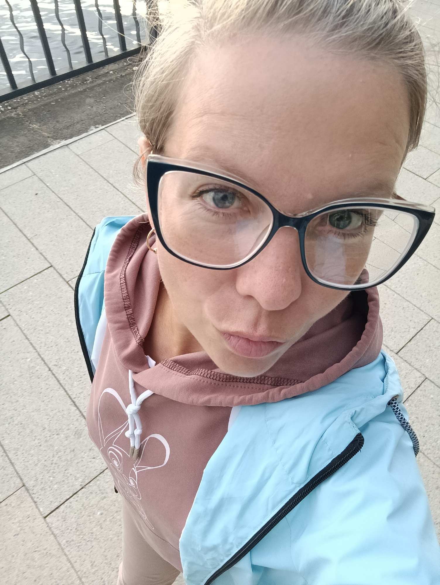 An outdoor, high-angle, medium shot captures a Caucasian woman with blonde hair pulled back and wearing glasses, a dusty rose hoodie, and a light blue jacket. She is looking up at the camera with a neutral expression. The background shows a paved walkway with a railing and water. The scene appears to be at the embankment of the Votkinskoye Reservoir in Chaikovsky, Russia, due to the characteristic railing and the presence of the reservoir.