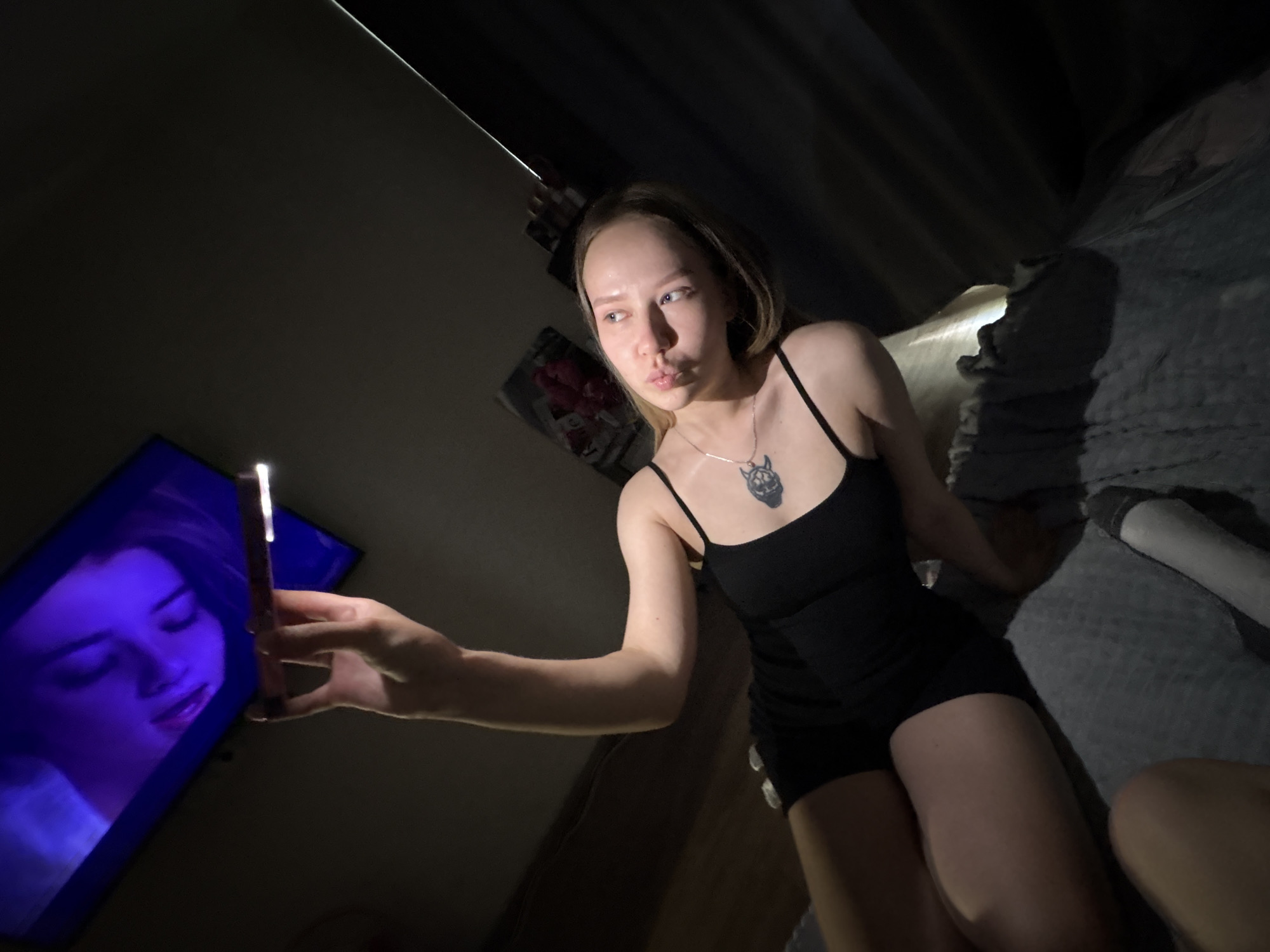 A young woman sits on a bed in a dimly lit room. She wears a black tank top and shorts, and a necklace. She is holding a smartphone in her hand, seemingly taking a selfie. A television screen is visible on the left, displaying an image of a woman. The room appears to be a bedroom. In the background, there is a covered object near the head of the bed, and a poster on the wall. The location is Chaikovsky, Russia.