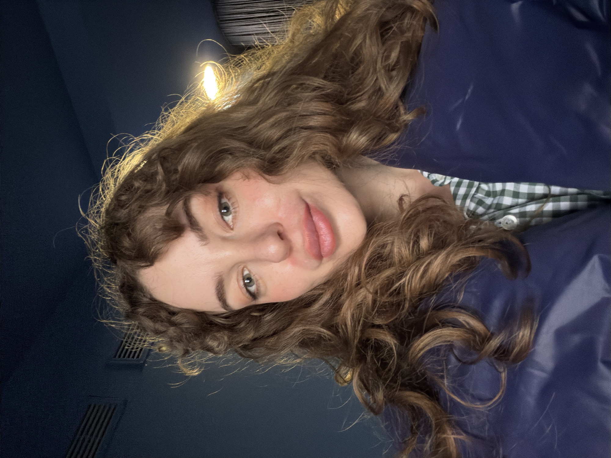 A close-up, downward-facing selfie shows a woman with light skin, full lips, and shoulder-length brown curly hair with wispy bangs. Her eyes are green. She is wearing a blue jacket and a collared shirt with a green and white checkered pattern. The background is a dark blue color with what appears to be a vent visible at the top left. A light source is above and slightly behind the woman. It is not possible to determine the location of the image to be in Moscow, Russia.