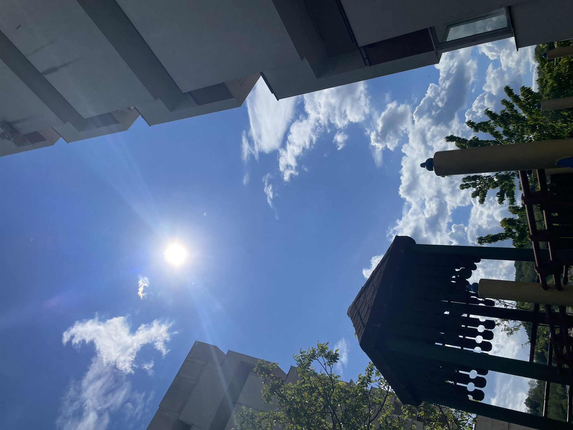 The image shows a view looking up towards the sky in Zlatograd, Bulgaria. A bright sun is visible, surrounded by a blue sky with scattered clouds. Buildings are situated on the left side of the frame. On the right side, part of a playground structure is visible, with green leaves from trees and bushes filling gaps in the background.