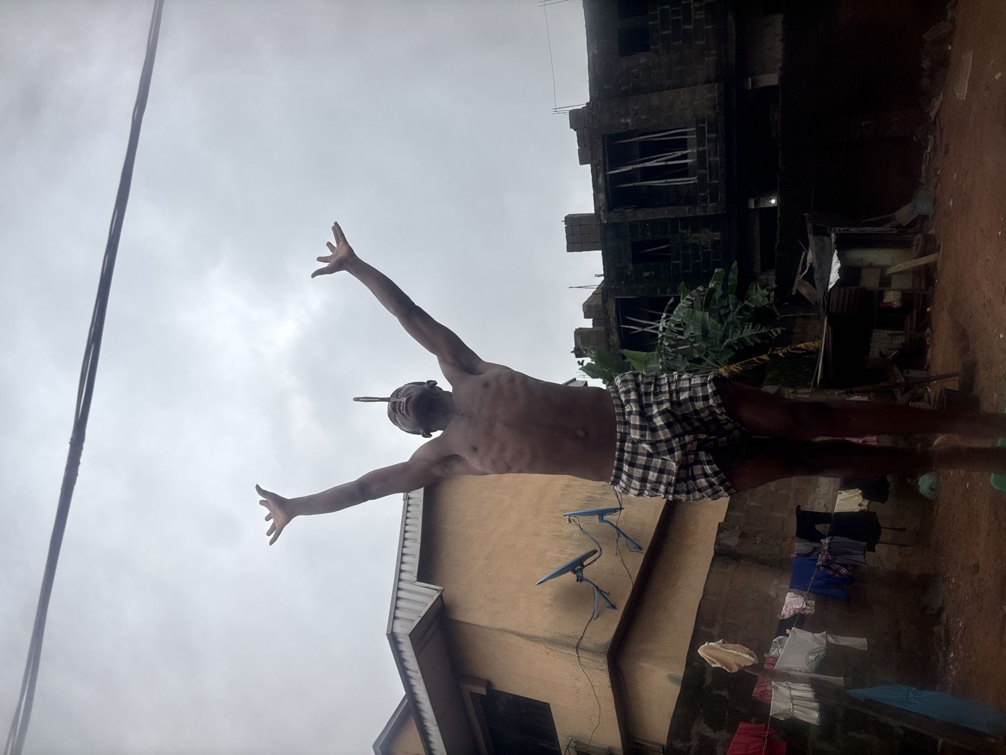 A young man is captured in a low-angle shot, standing outdoors with his arms outstretched towards the sky in Nkpor, Nigeria. He is wearing plaid shorts and appears to be balancing a thin object on his nose. Behind him, a two-story building with a light brown facade and a corrugated metal roof is visible, equipped with two satellite dishes. To the right, a partially constructed dark brick building with visible wooden supports stands against an overcast sky. In the foreground, laundry hangs on a clothesline strung between poles. The ground is covered in reddish dirt, with various scattered objects including what appears to be a blue plastic bucket and some debris. The overall lighting suggests a cloudy or dim daytime environment.