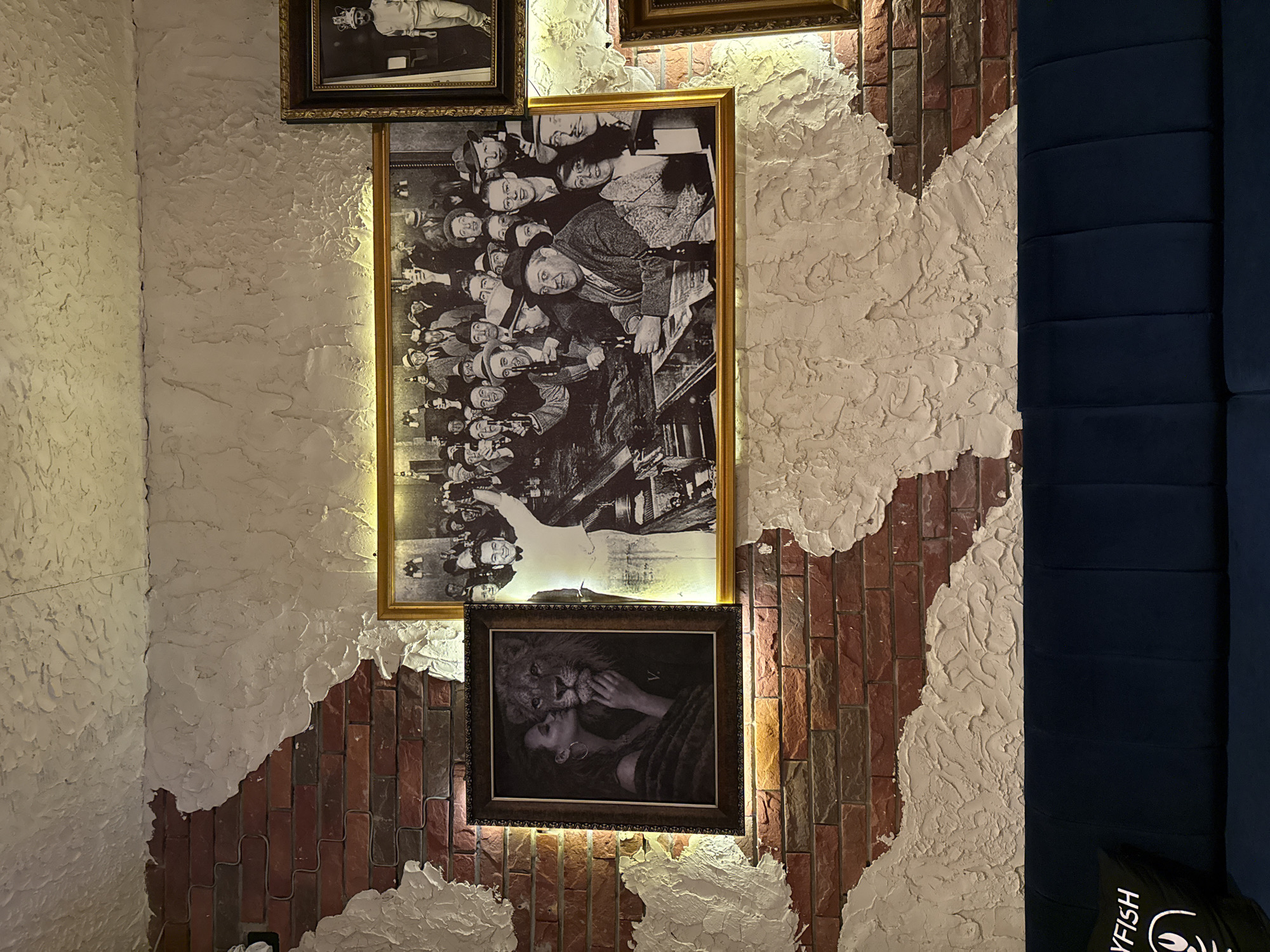 The media file captures an interior wall decorated with framed black and white artworks, illuminated by warm spotlights, likely in a restaurant or lounge in Kuta, Indonesia. The wall features a distinctive design with heavily textured, off-white plaster that appears to be partially "peeling away" or intentionally applied to reveal sections of underlying red and brown brickwork.
Three framed pictures are prominently displayed. The largest, centrally located, is a gold-framed black and white image of a dense crowd of people, many smiling and looking upwards, suggestive of an audience at an event. This picture is rotated 90 degrees clockwise. Below it, a dark-framed black and white photograph depicts an intimate scene of a woman looking up at a lion, who gently places a paw on her face as she touches its snout. A smaller, dark-framed black and white image in the top-left shows a person, possibly a man in formal attire, in an ambiguous pose.
To the right, a dark blue padded wall or seating area contrasts with the textured wall. A black bag with white text that reads "YFISH" and a circular logo with a stylized face is visible resting on this blue surface. The lighting creates a cozy and dramatic ambiance, highlighting the textures of the wall and the artworks.