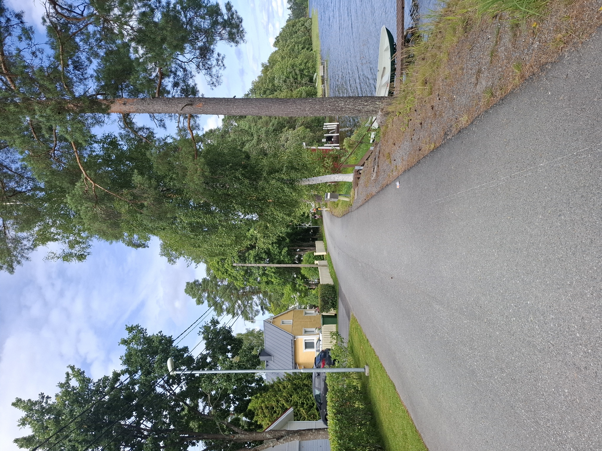 This image captures a tranquil scene in Mikkeli, Finland, likely on a summer day. The perspective is from a paved road that curves to the left, bordered by a grassy lawn and trees on the left and the calm waters of a lake on the right.
On the left side of the road, a bright yellow house with a grey roof is visible, set back from the road and surrounded by green hedges. A dark-colored car is parked in front of a white house with a garage. A lamppost stands on the left side of the road near the yellow house.
The right side of the image is dominated by the lake, with a wooden pier extending into the water. A light-colored boat with a green trim is resting on the pier. A tall pine tree stands prominently in the foreground on the right, with a dense canopy of green foliage. Further back along the lakeshore, more trees create a lush, green backdrop. The sky above is blue with scattered white clouds, suggesting fair weather. The overall atmosphere is one of peace and natural beauty.