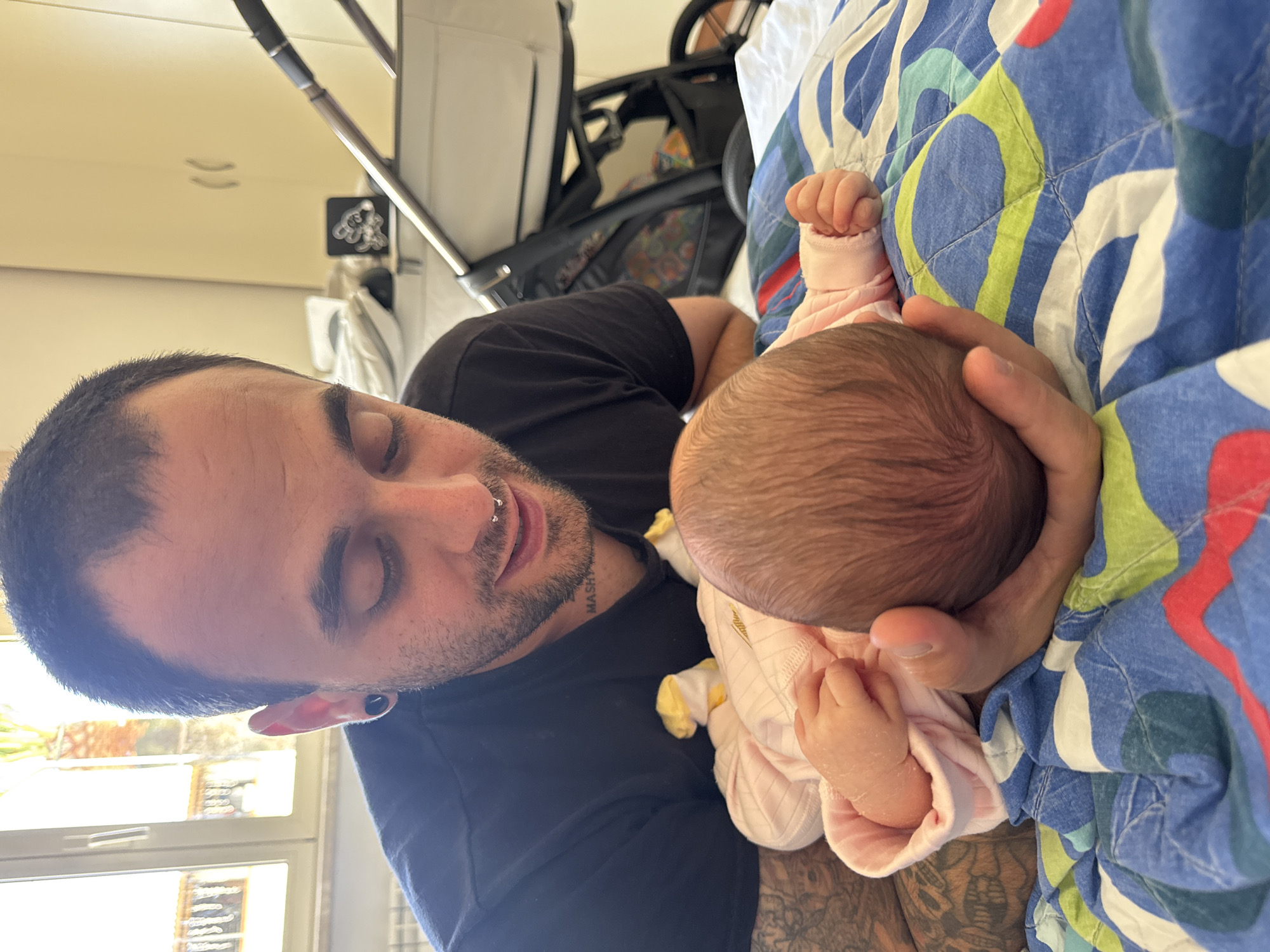 A medium shot depicts a fair-skinned man with short dark hair and a beard, facing the camera at a slight angle, holding a baby with dark hair. The man is wearing a black t-shirt and has visible tattoos on his arm. He has a piercing in his septum and an ear piercing. He gazes down at the infant. The baby is wearing a light pink outfit. They are resting on a quilt with a blue, green, white, and red pattern. Behind them is a baby stroller and a window. The location is implied to be Aprilia, Italy, by external information, although this cannot be directly confirmed from the image.